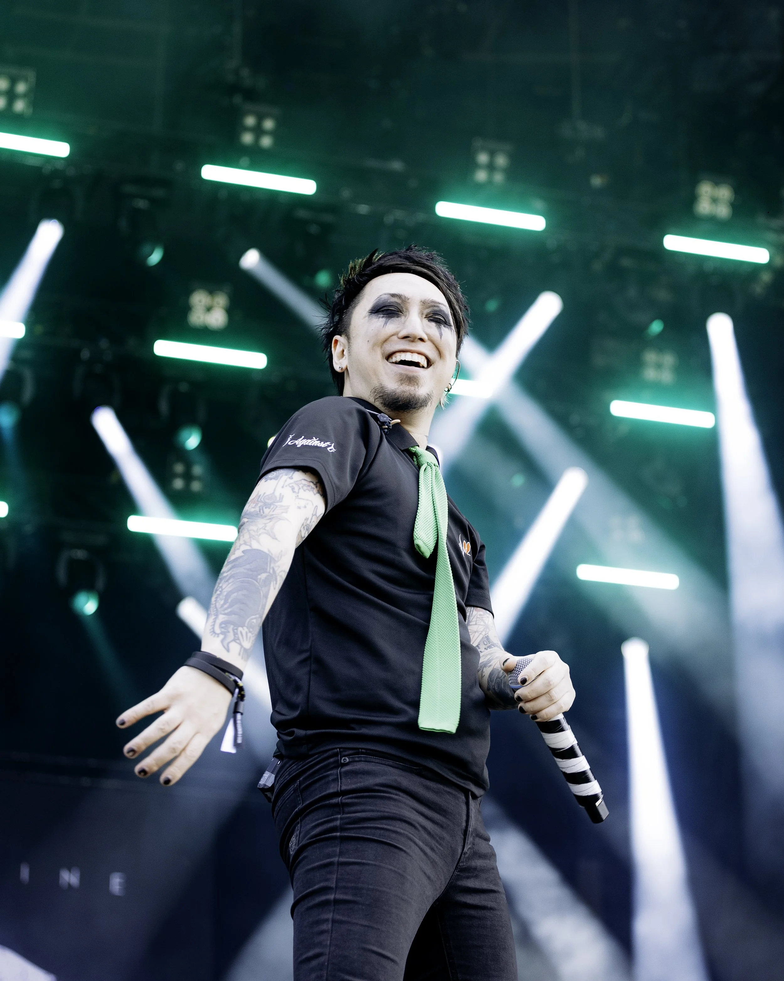 A male performer with tattoos on his arms singing into a microphone on stage with green and white stage lights in the background.