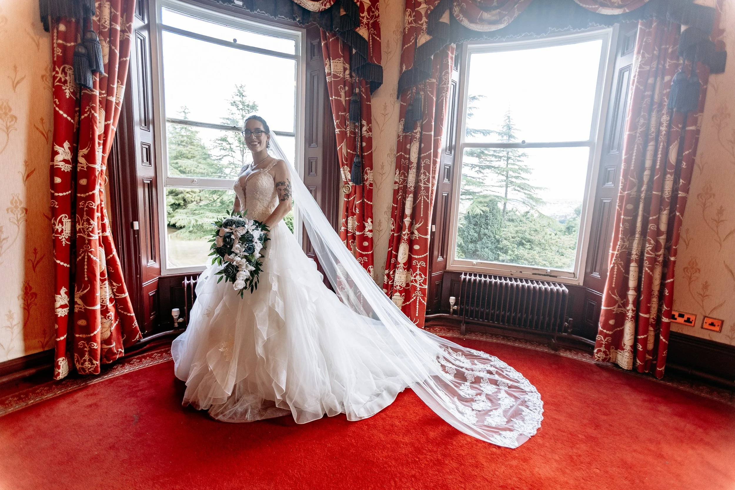 A bride in a wedding dress holding a bouquet, standing by large windows in a historic room with red patterned curtains and carpet, and scenic greenery outside.