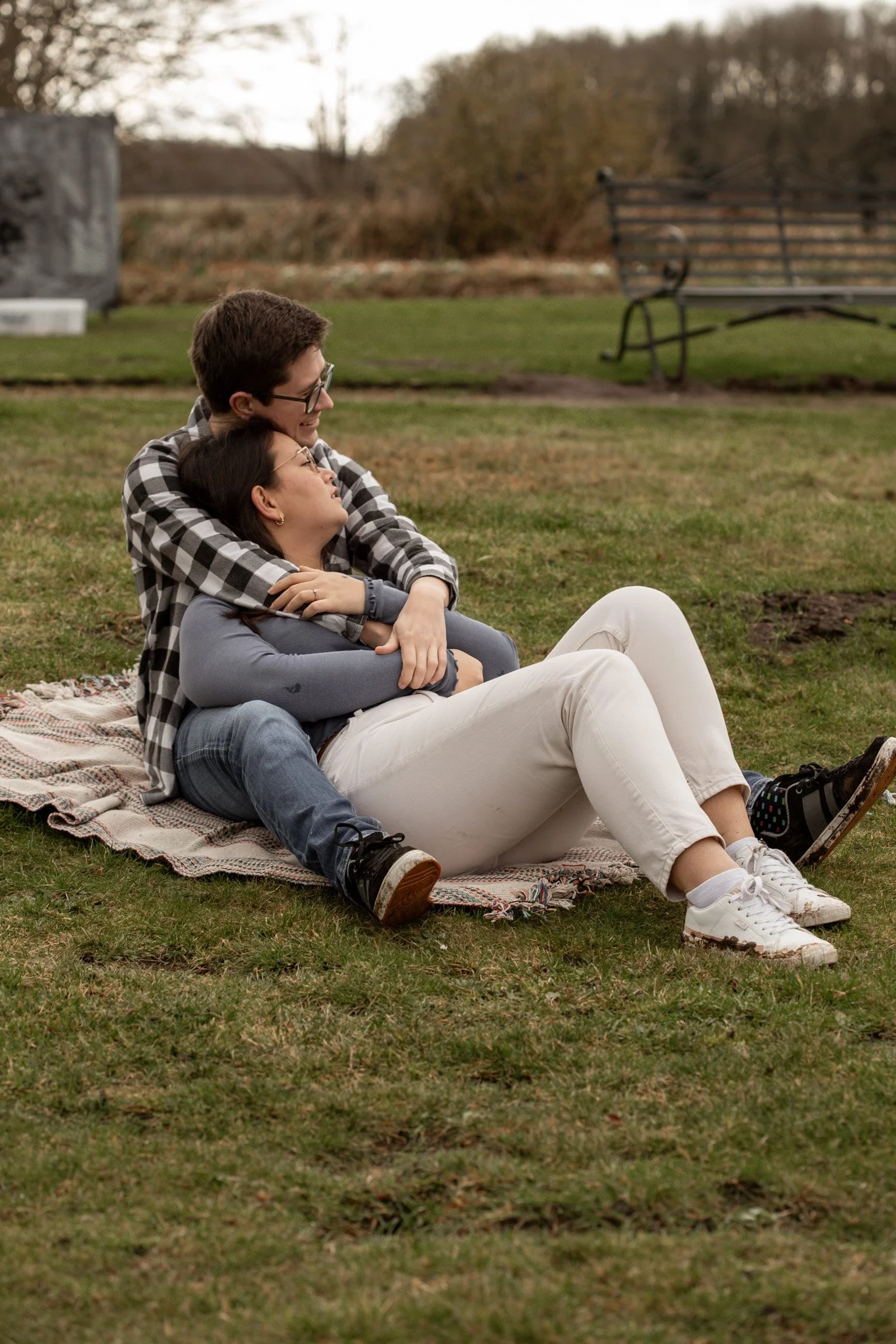 Two people sitting on a blanket in a park, embracing each other, with trees and a bench in the background.