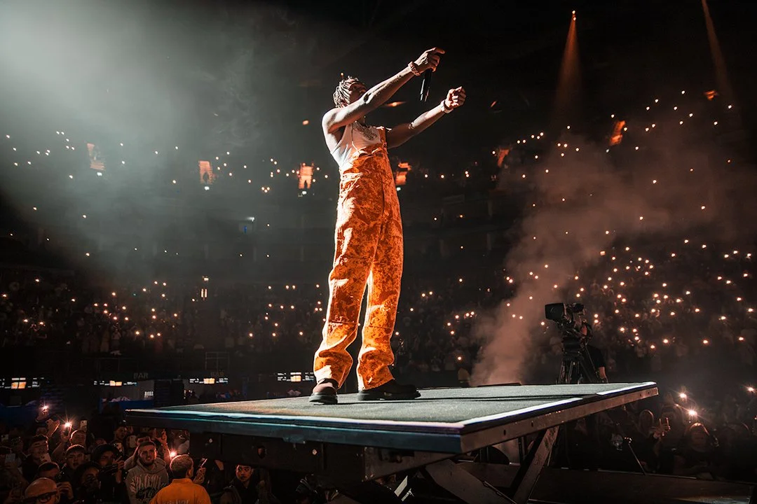Performer in orange overalls on stage with microphone at a concert with audience holding lights.