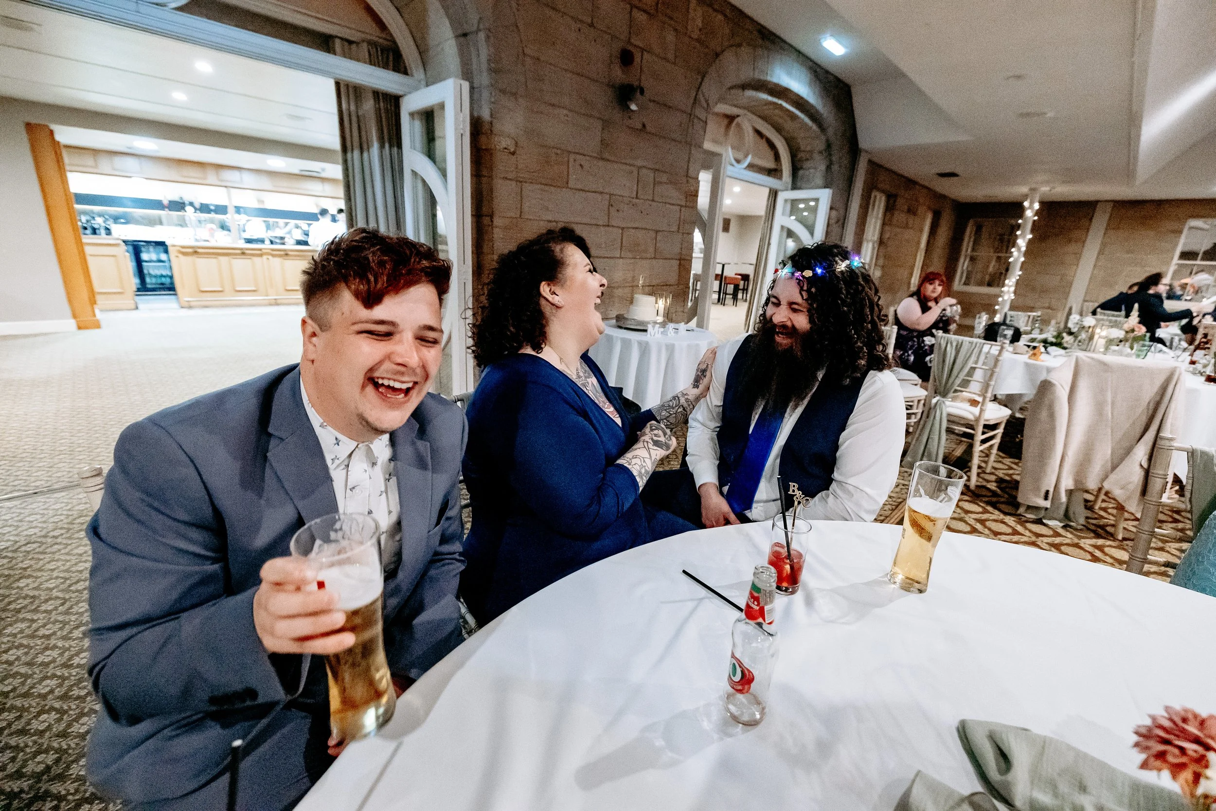 Four people at a wedding reception sitting around a table, three of them are laughing and smiling, with drinks in front of them.