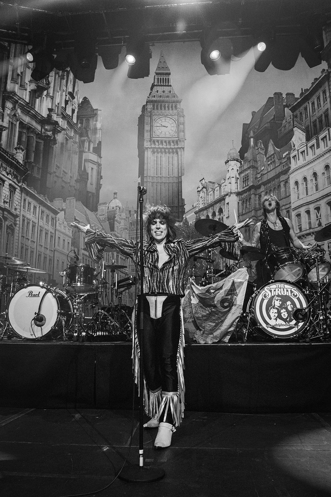Black and white photo of a musician on stage, arms outstretched, with a cityscape backdrop featuring Big Ben. Drum kit with "The Struts" logo in the background.