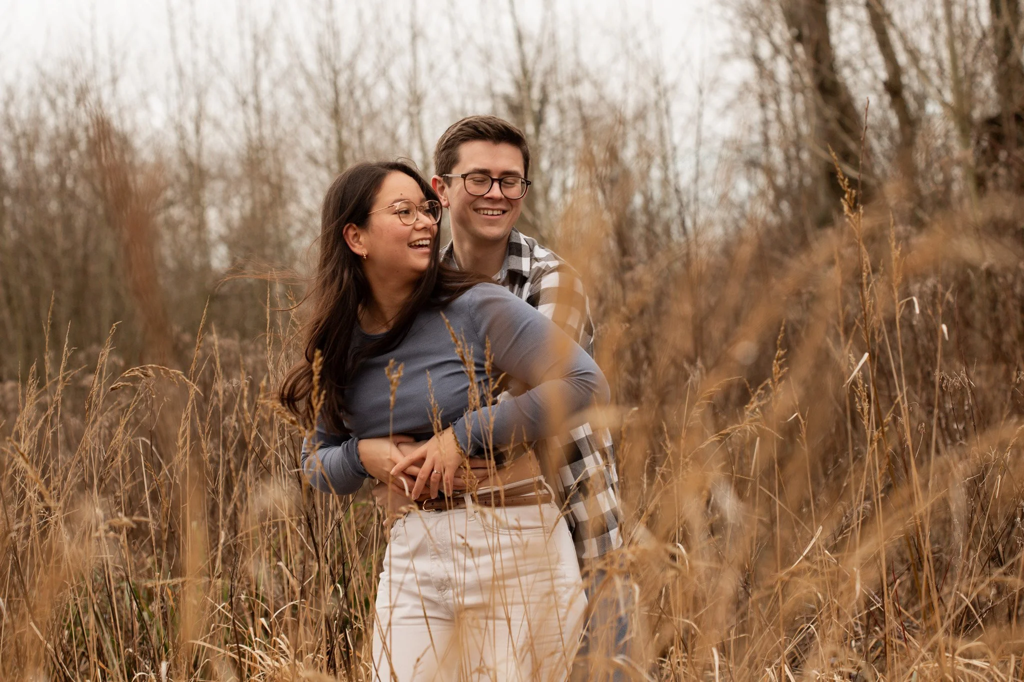 A smiling young couple standing in a field of tall, dry grass with trees in the background, embracing each other.
