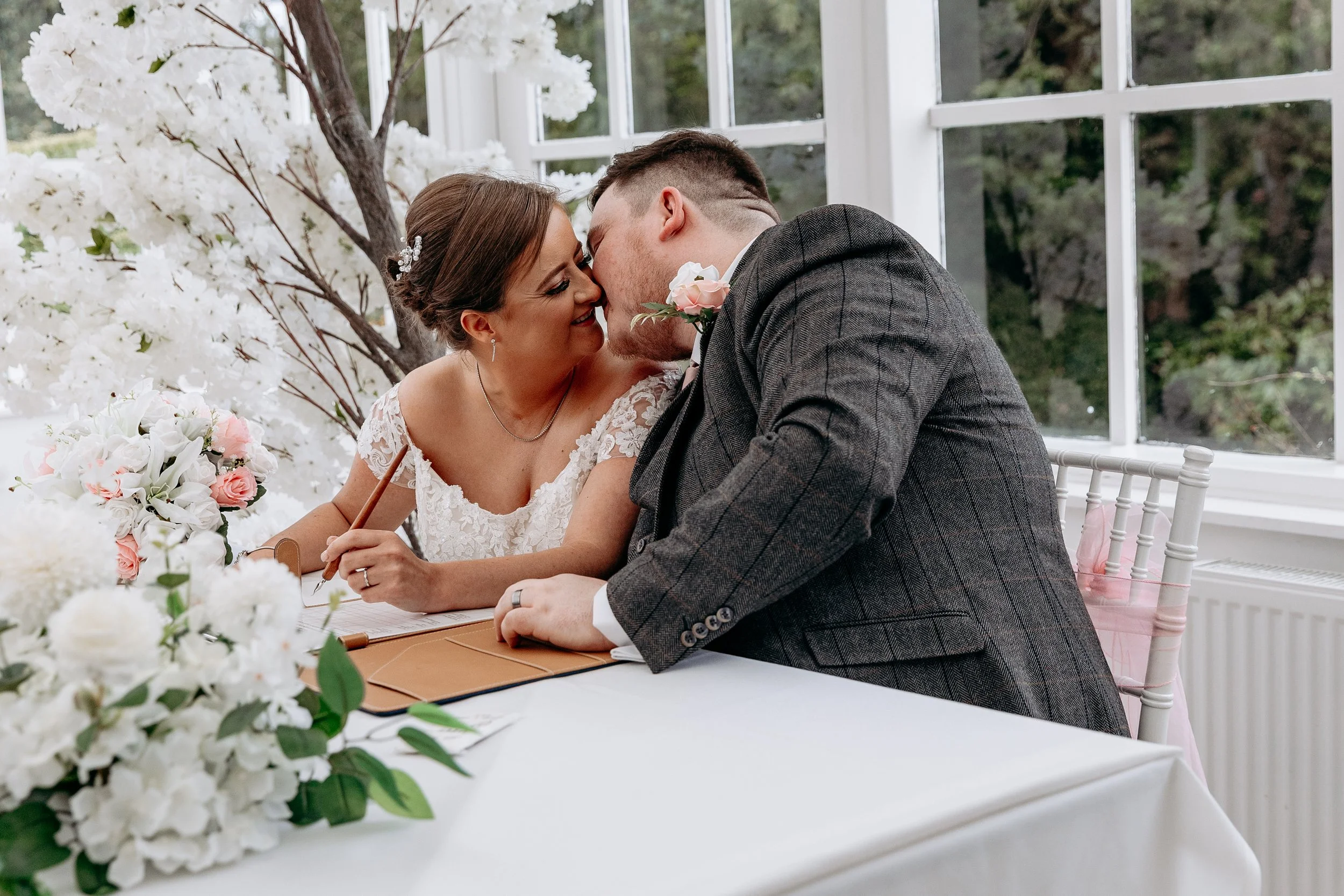 A bride and groom kiss at a wedding ceremony, with flowers and a white floral backdrop in the background.