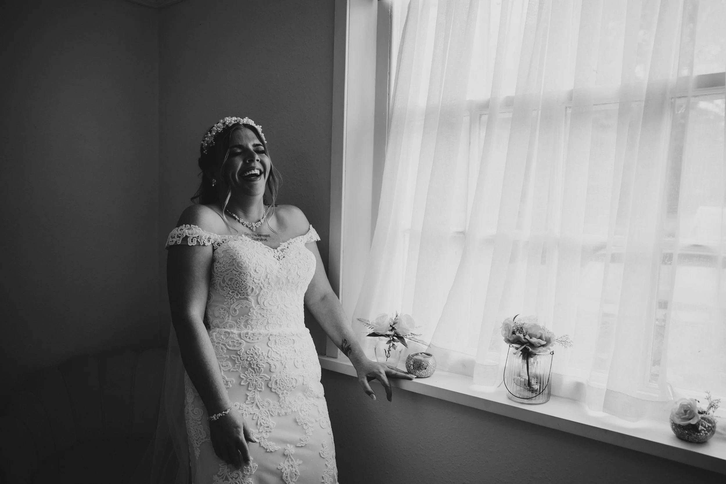 A bride in a lace wedding dress smiling and laughing inside a room, standing near a window with sheer curtains and small flower arrangements on the windowsill.