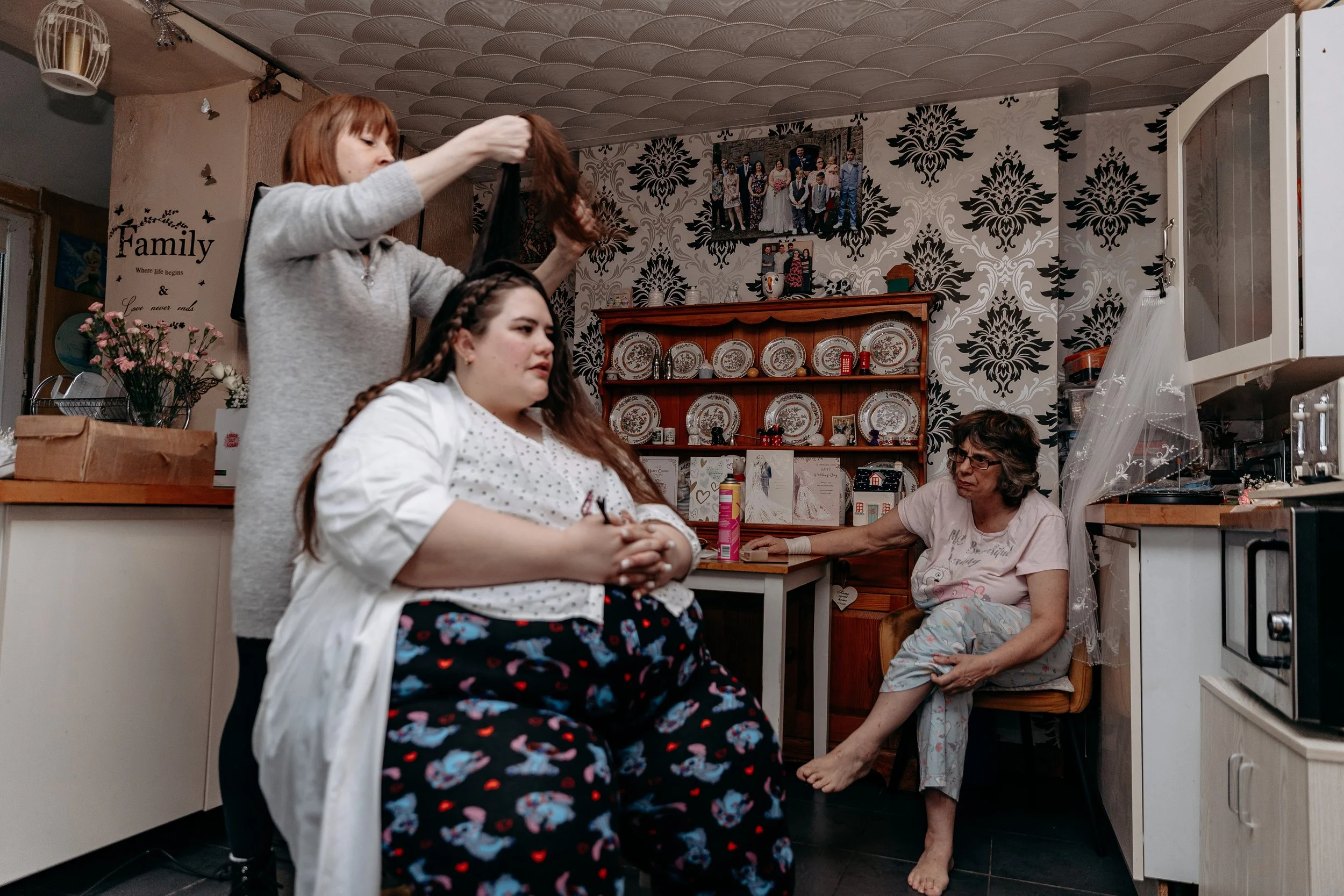A woman sitting in a living room being attended to by two other women, with a third woman sitting nearby. The scene depicts a casual indoor setting with a wooden shelf displaying plates and decorations, floral wallpaper, and personal photographs on t