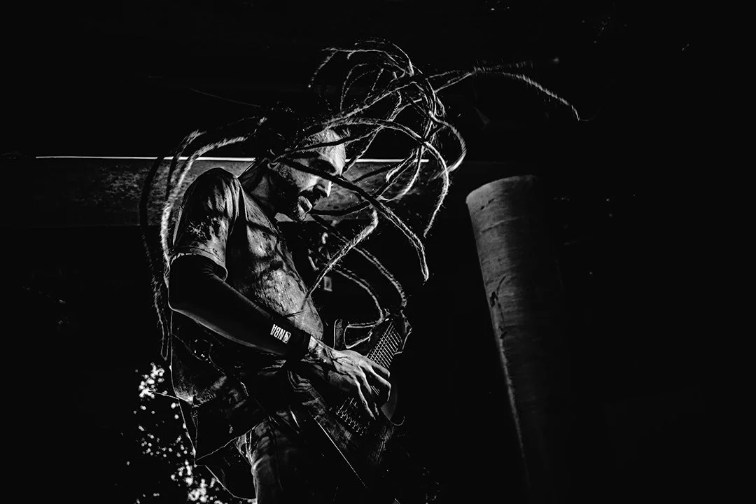 A black and white photo of a musician with dreadlocks playing an electric guitar, standing on a dark stage.