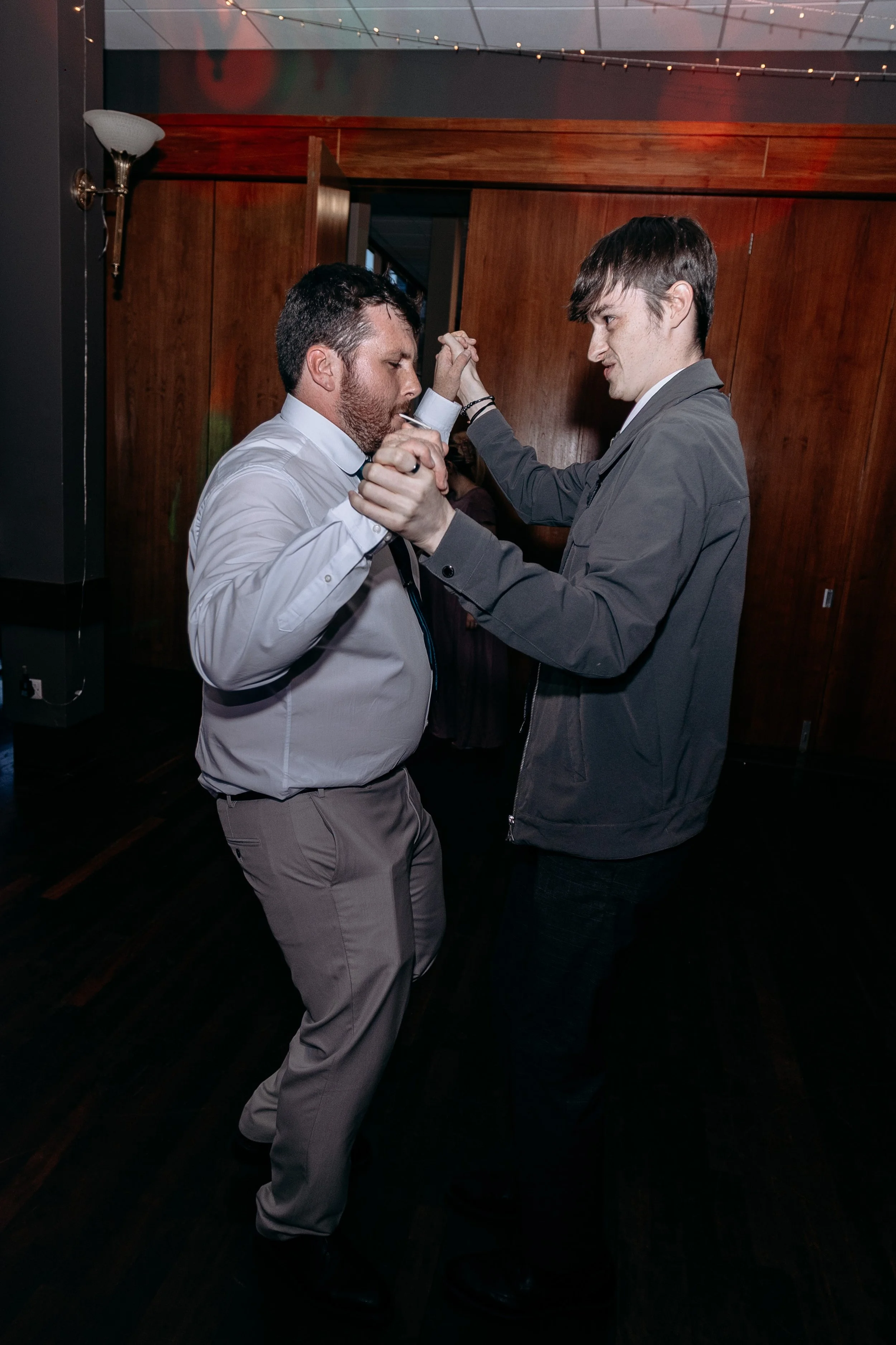 Two men dancing closely at an indoor party with wooden walls and string lights overhead.