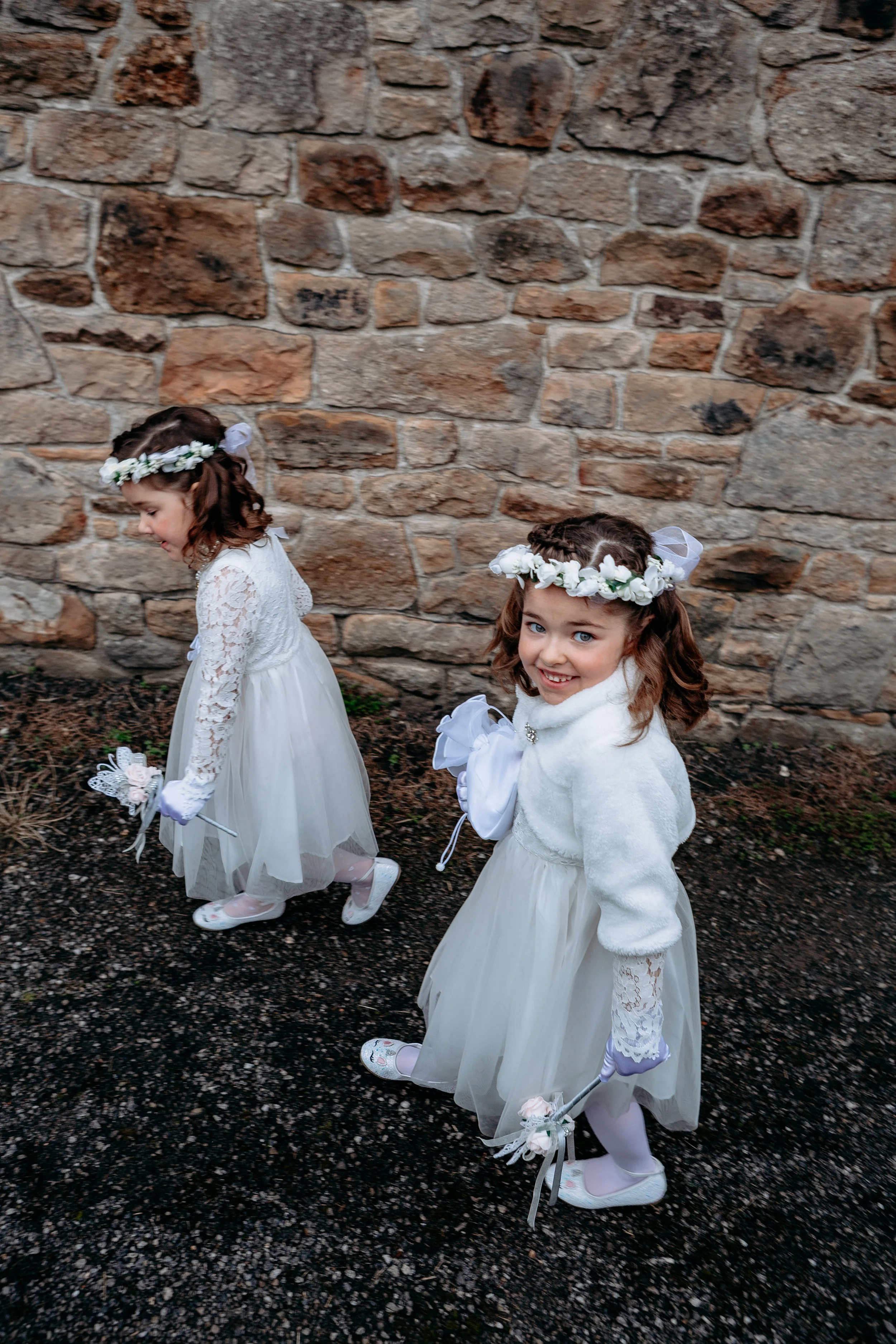 Two young girls dressed in white flower girl dresses walking outdoors near a stone wall, each holding a small bouquet, wearing floral headbands and smiling.