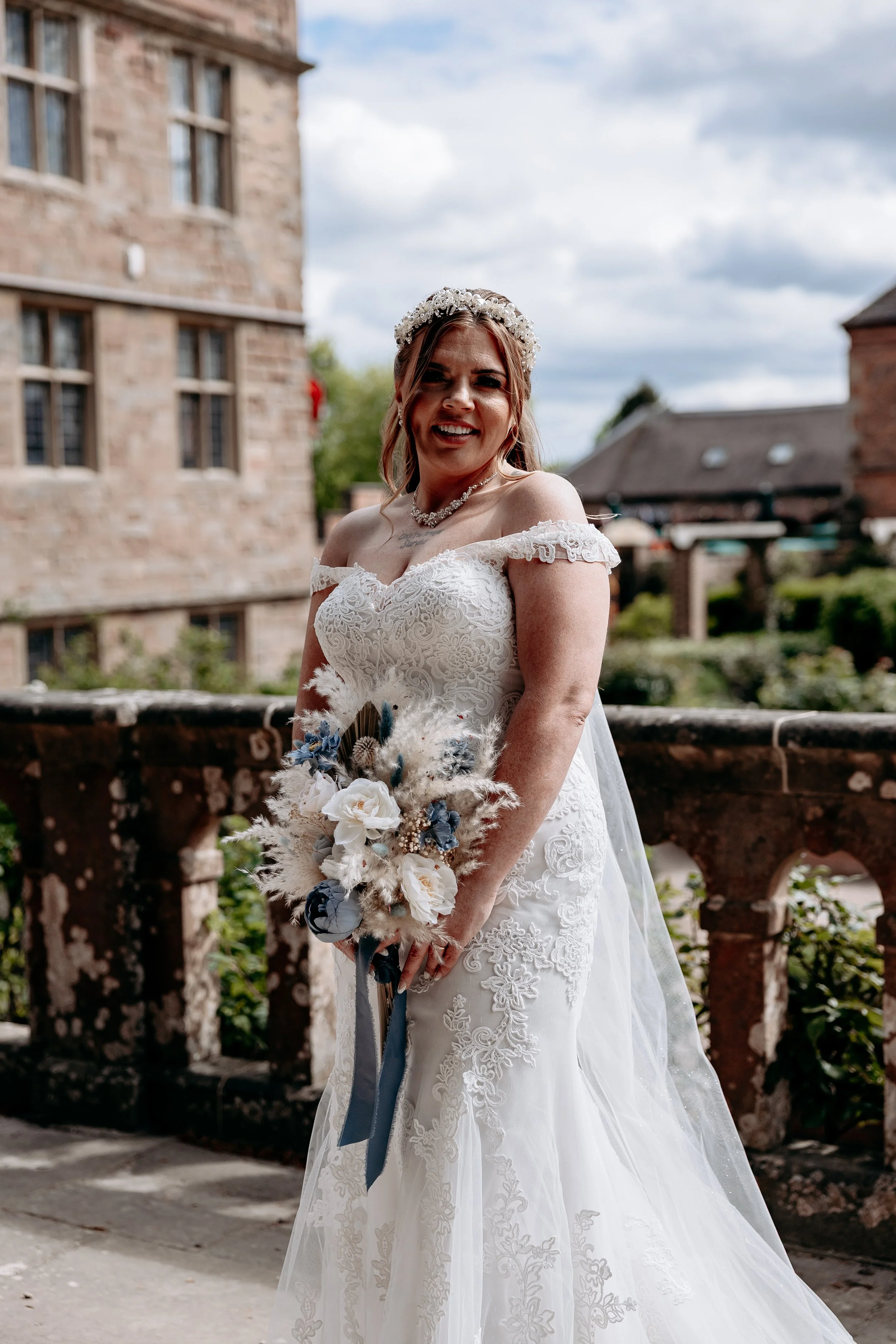 A smiling woman in a white lace wedding dress holding a bouquet of white and blue flowers, standing outdoors on a stone balcony with historic stone buildings and a cloudy sky in the background.