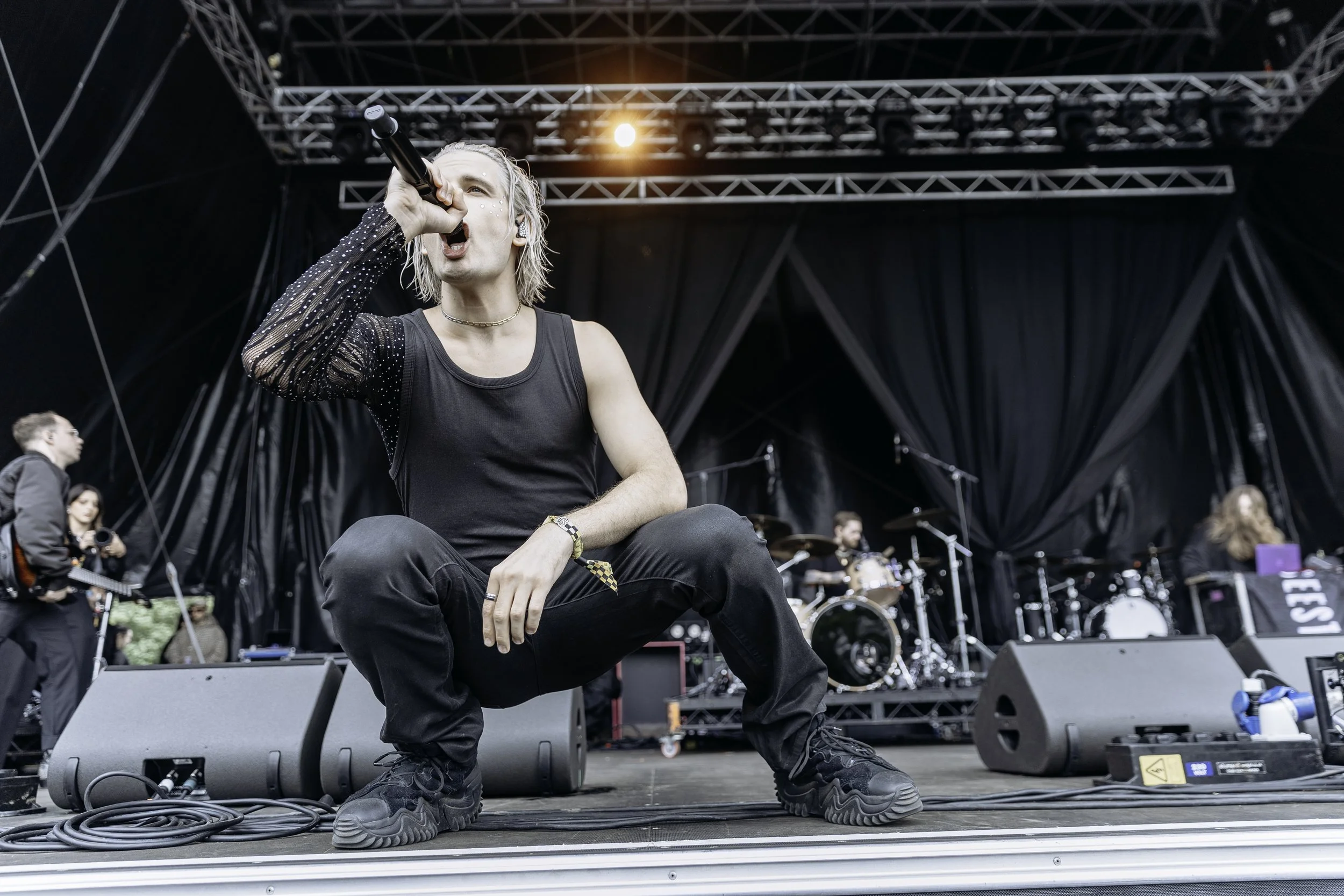 Male singer crouching and singing into a microphone on a stage, surrounded by musicians and instruments, during a live music performance.