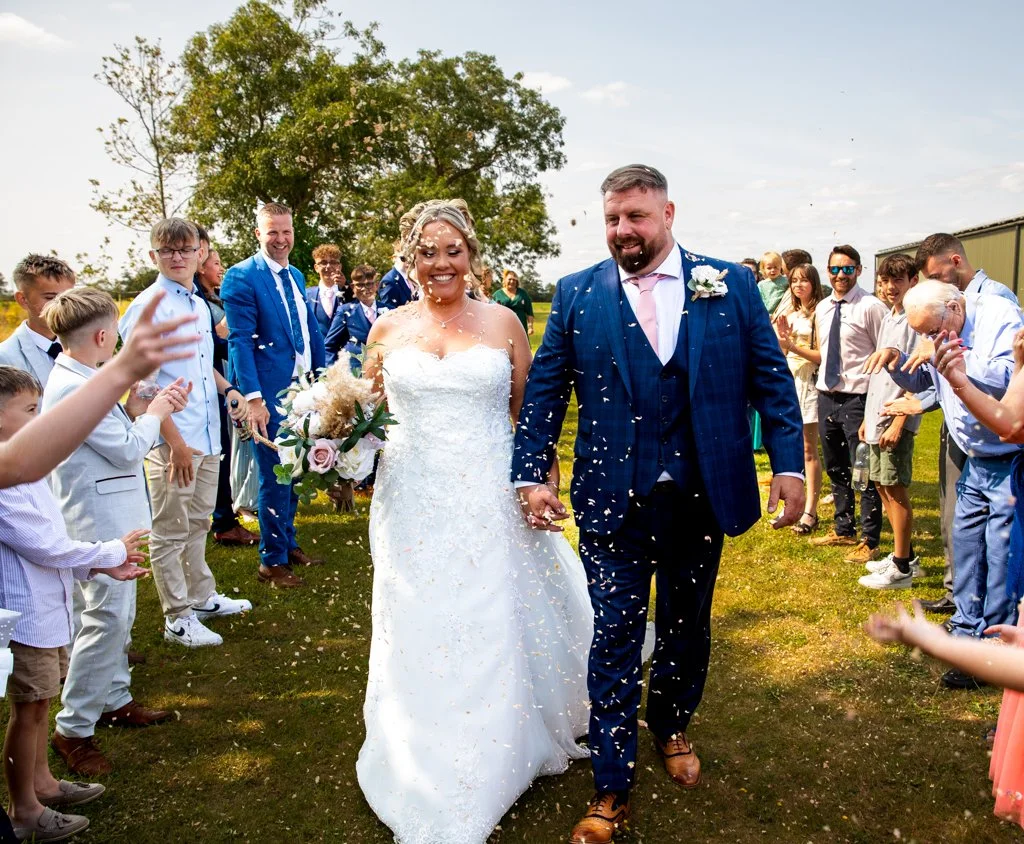 Bride and groom walking through a crowd of guests during their outdoor wedding celebration, with guests throwing confetti and a large tree in the background.