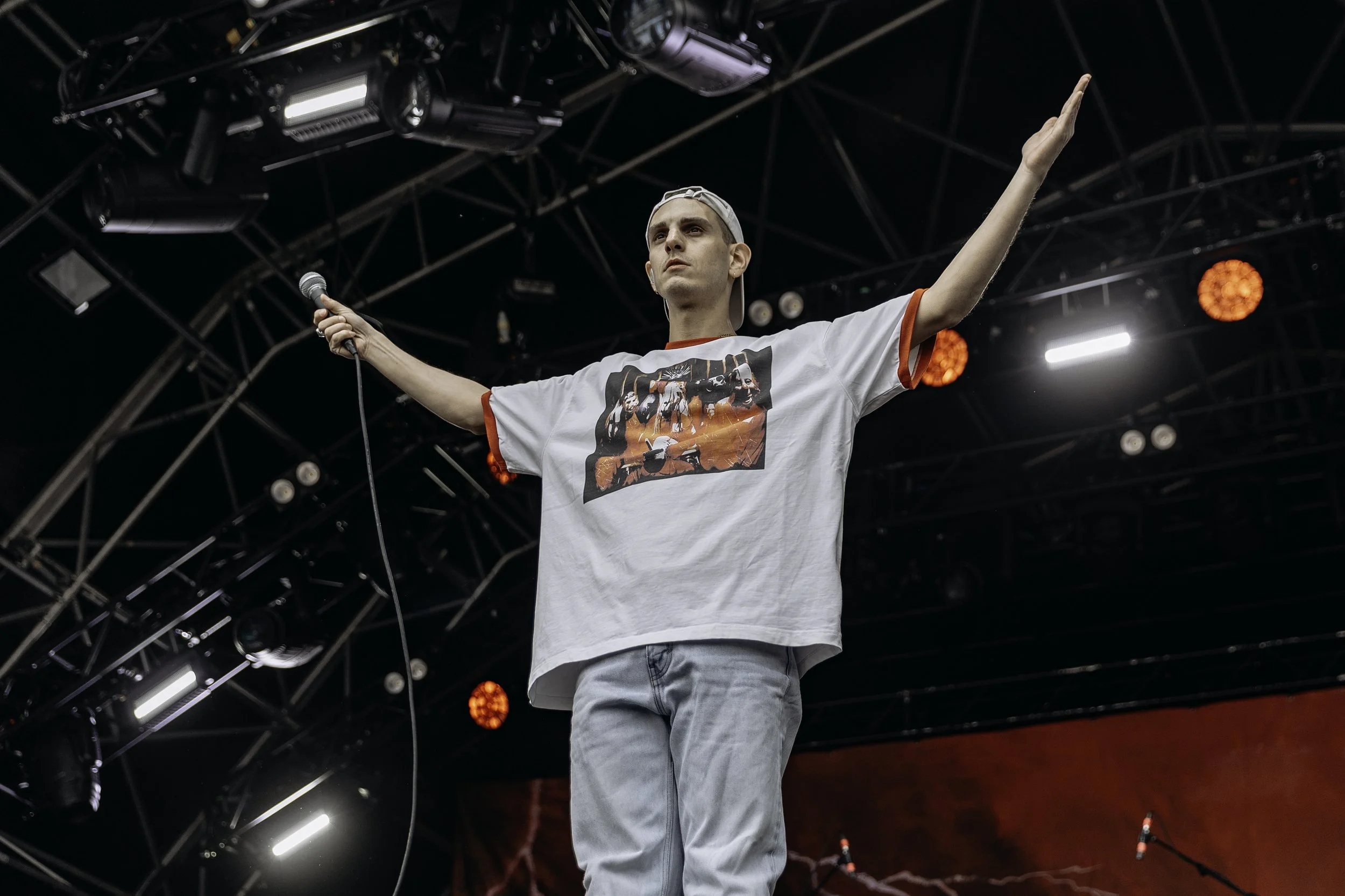 A man with short hair wearing a white T-shirt with a graphic print and light jeans, holding a microphone in his right hand, standing on a stage with arms extended, and a black security cap worn backwards. The stage has black metal framework, and red 