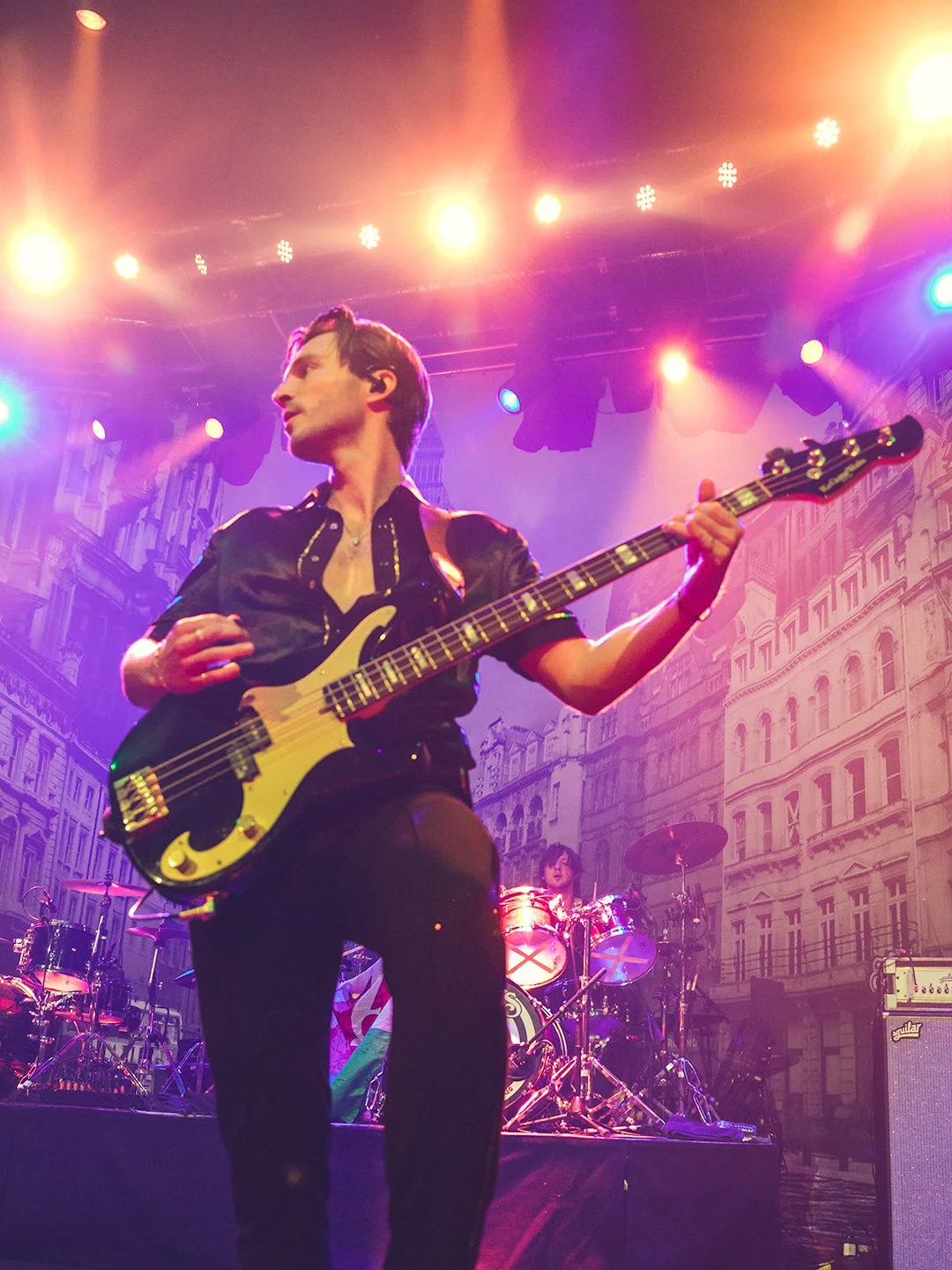A musician playing a bass guitar on stage with bright lights and a building backdrop. Another musician is visible playing drums in the background.