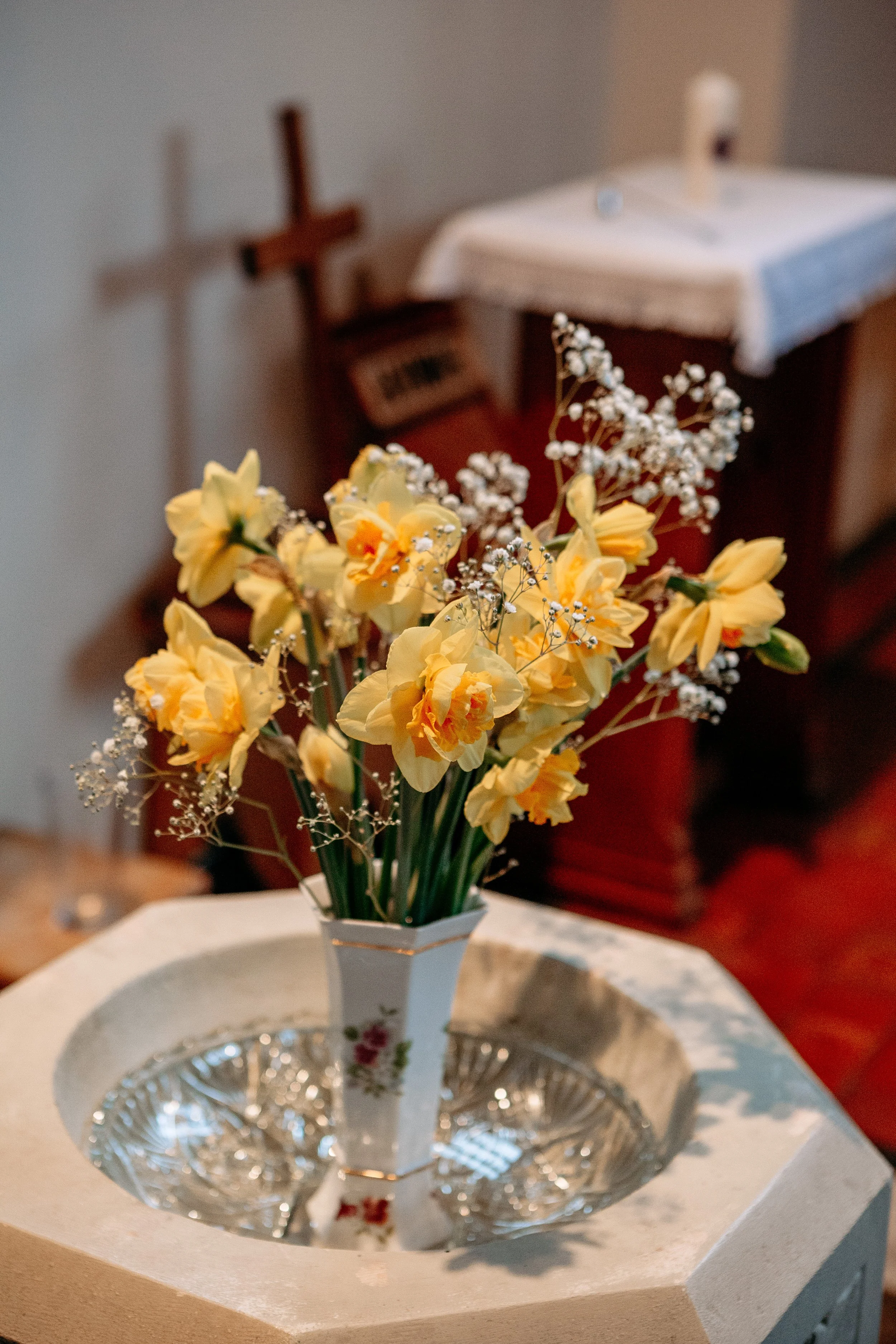 A vase with yellow daffodils and white baby's breath on a white table with a glass bowl, with a cross and religious items in the background.