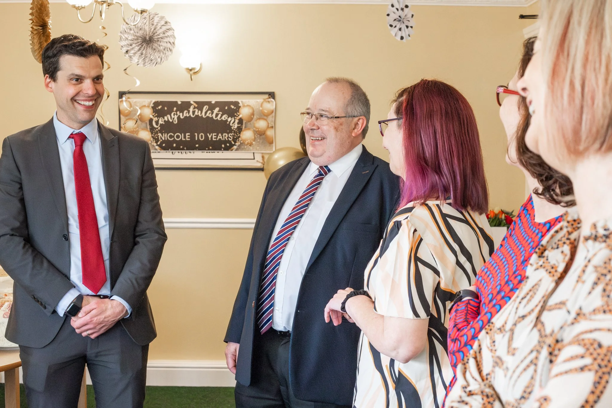 A group of five people, including a man in a gray suit with a red tie, smiling and talking at a celebration. Behind them is a decorated wall with a sign that says 'Congratulations! Nicole 10 Years' and gold and black decorations.