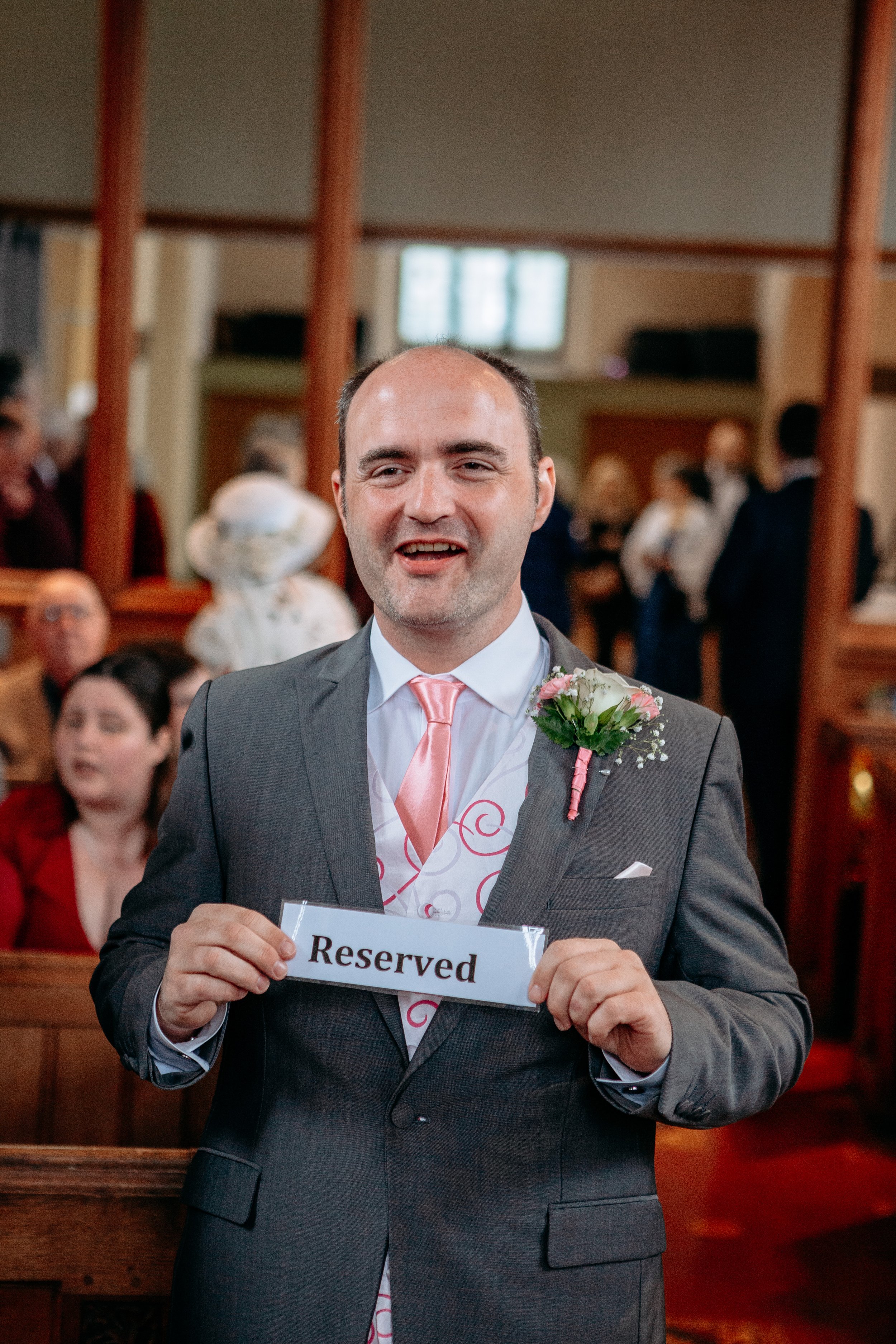 A man in a suit and a pink tie holding a reserved sign inside a church, with people in the background, during a wedding or event.