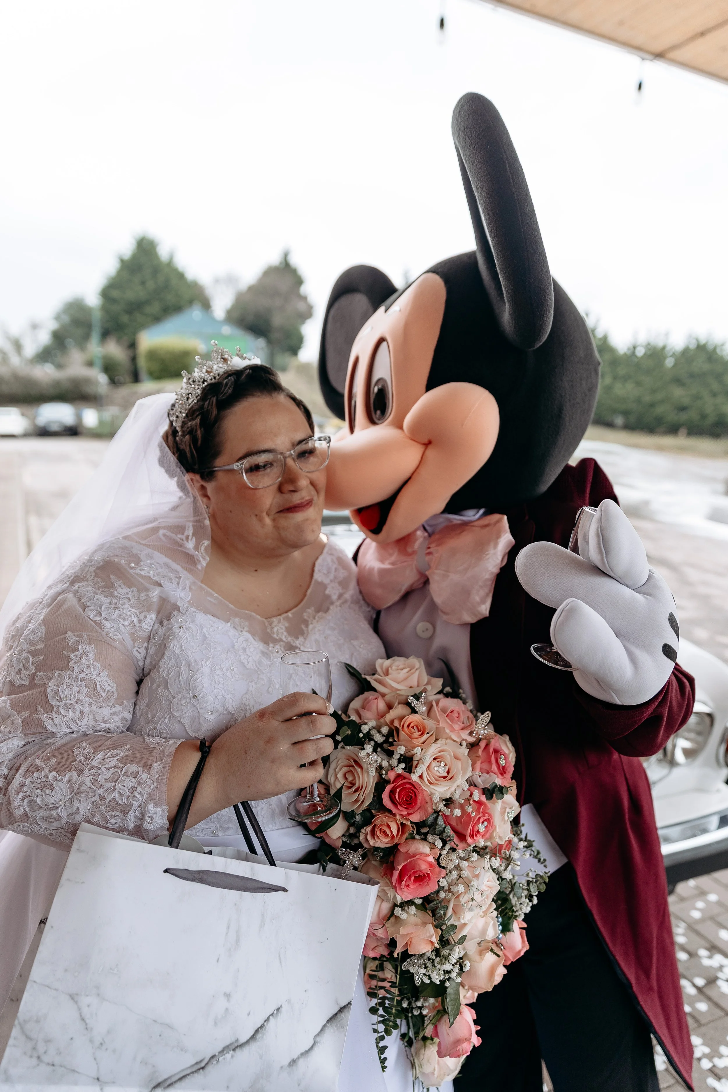 A bride in a wedding dress holding a bouquet of pink roses and a glass, with a person dressed as Mickey Mouse giving her a hug.