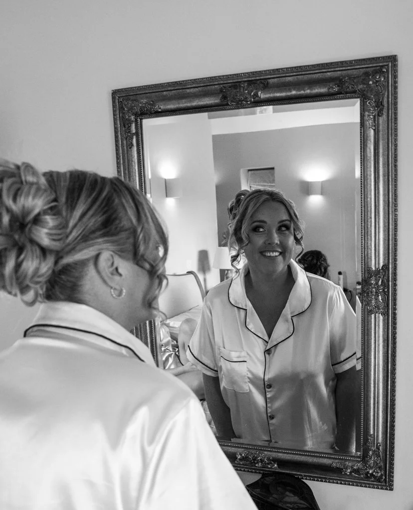 A woman smiling at her reflection in a decorative mirror. She is wearing satin pajamas and has styled, curled hair.