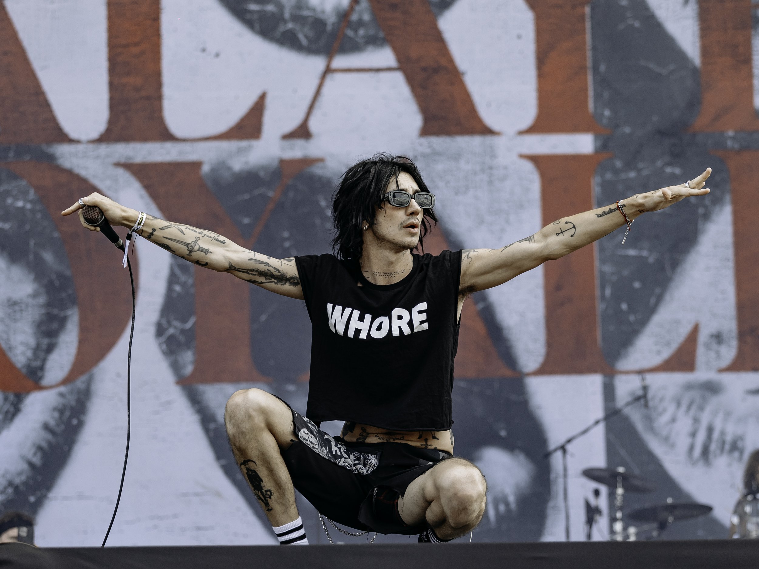 A male singer with tattoos, wearing sunglasses and a black crop top with the word 'WHORE' on it, performing with one arm extended while holding a microphone, on stage with a large backdrop.
