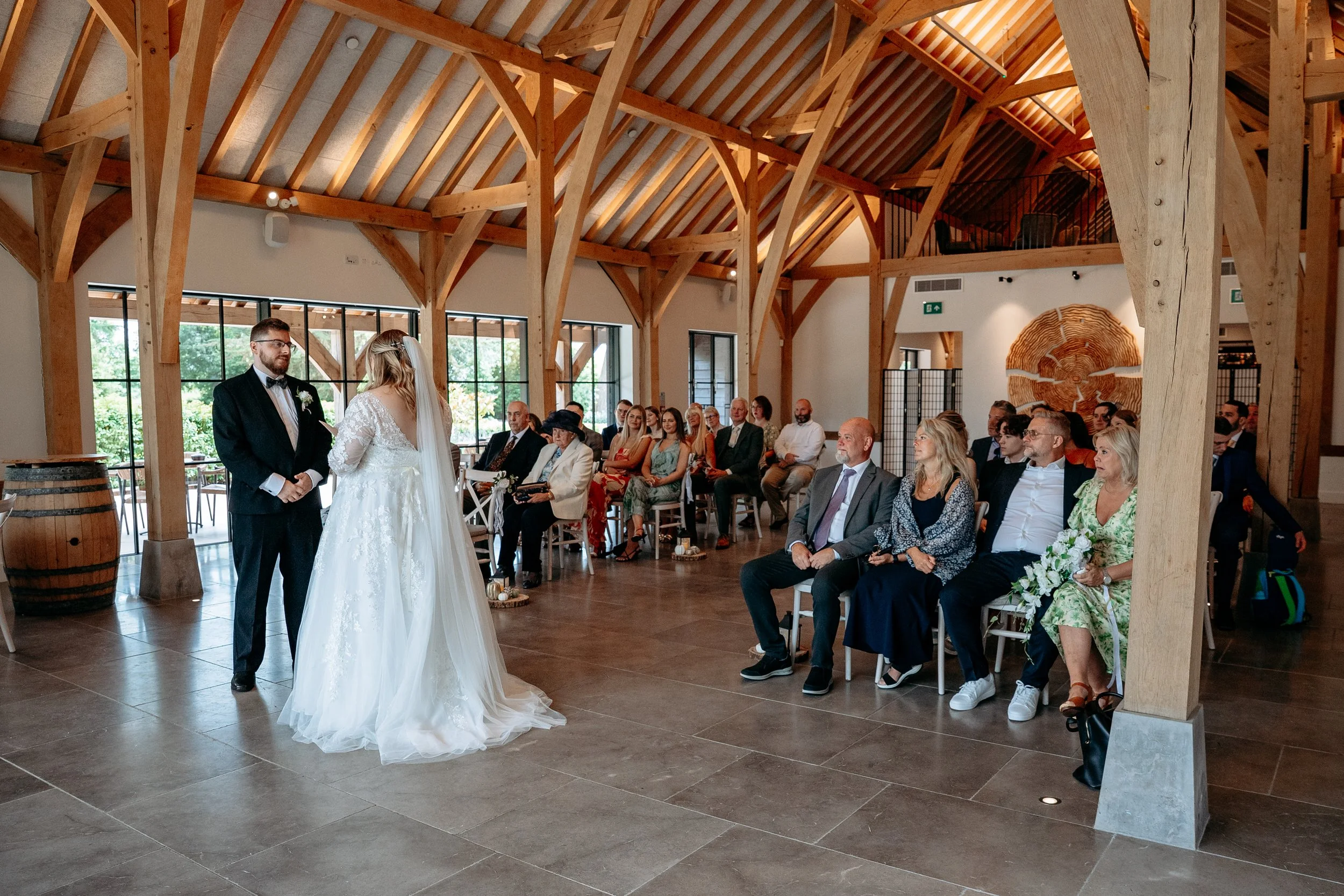 Indoor wedding ceremony with the bride and groom standing in front of seated guests in a wooden-beamed venue.