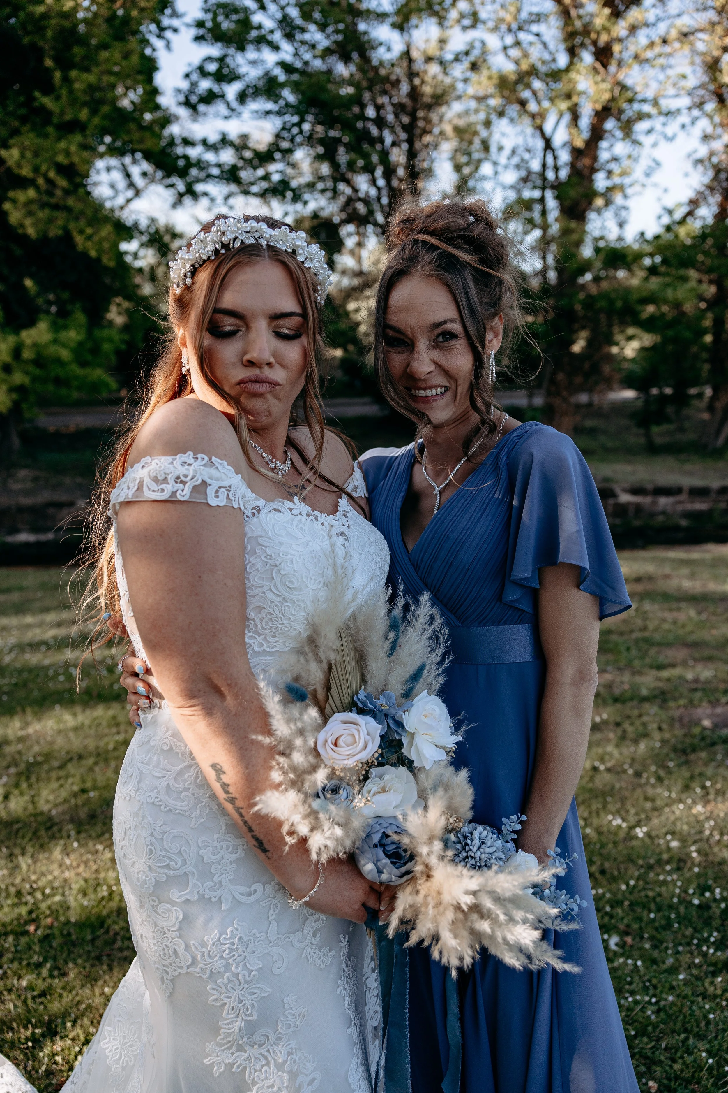 A bride in a white lace wedding dress holding a bouquet of white and blue flowers and pampas grass standing outdoors with a woman in a blue dress, both smiling and posing for the camera, surrounded by trees.