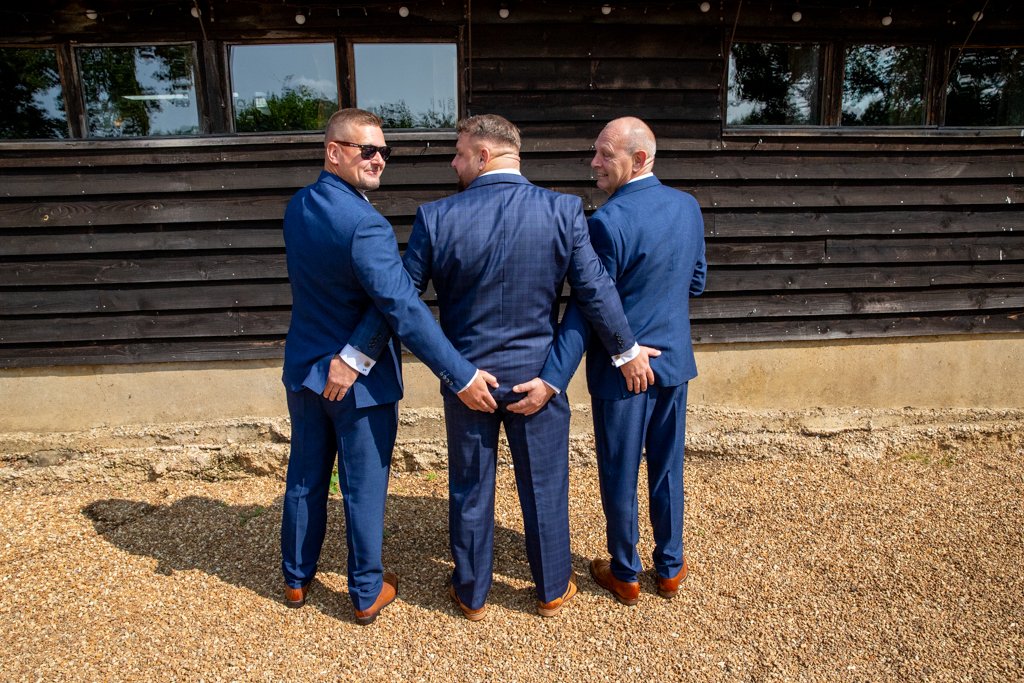 Three men in blue suits standing outside in front of a dark wooden building, holding hands behind their backs, with one man in the middle facing away and the other two facing sideways and smiling.