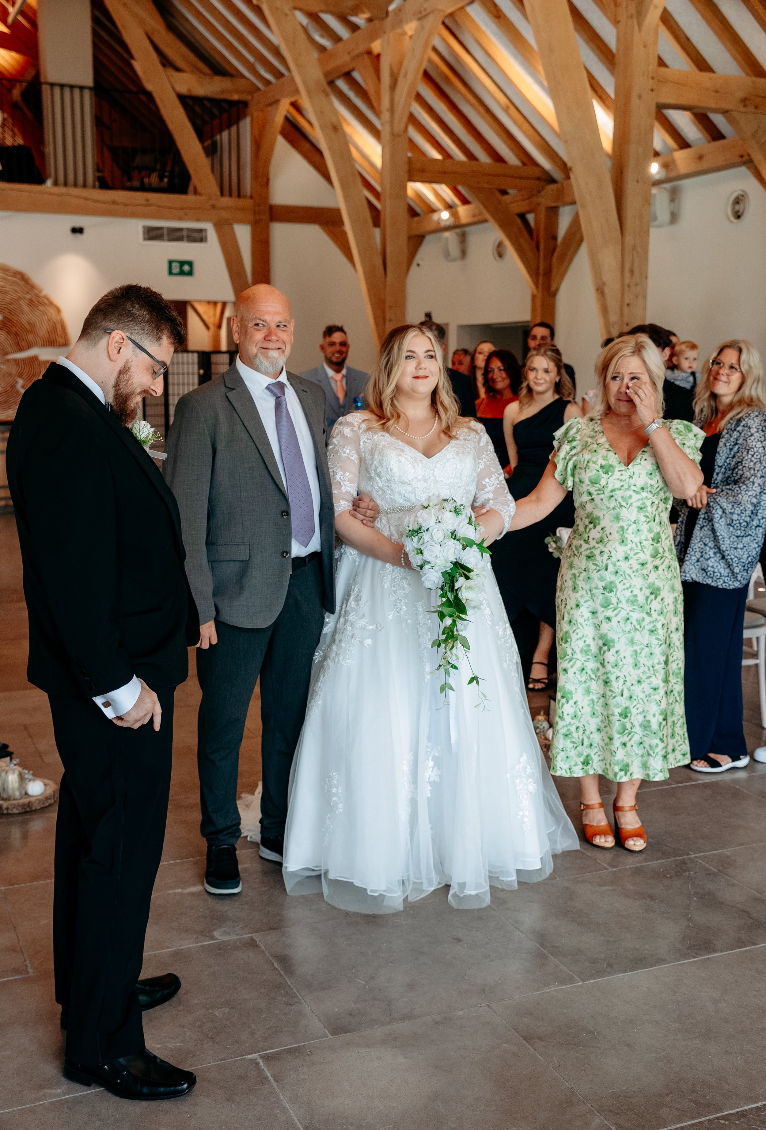 A bride in a white gown holding a bouquet stands arm-in-arm with a man in a gray suit inside a wooden-beamed venue. A woman in a green floral dress stands next to them, wiping her eye. A man in a black suit is looking down at the floor. A group of pe
