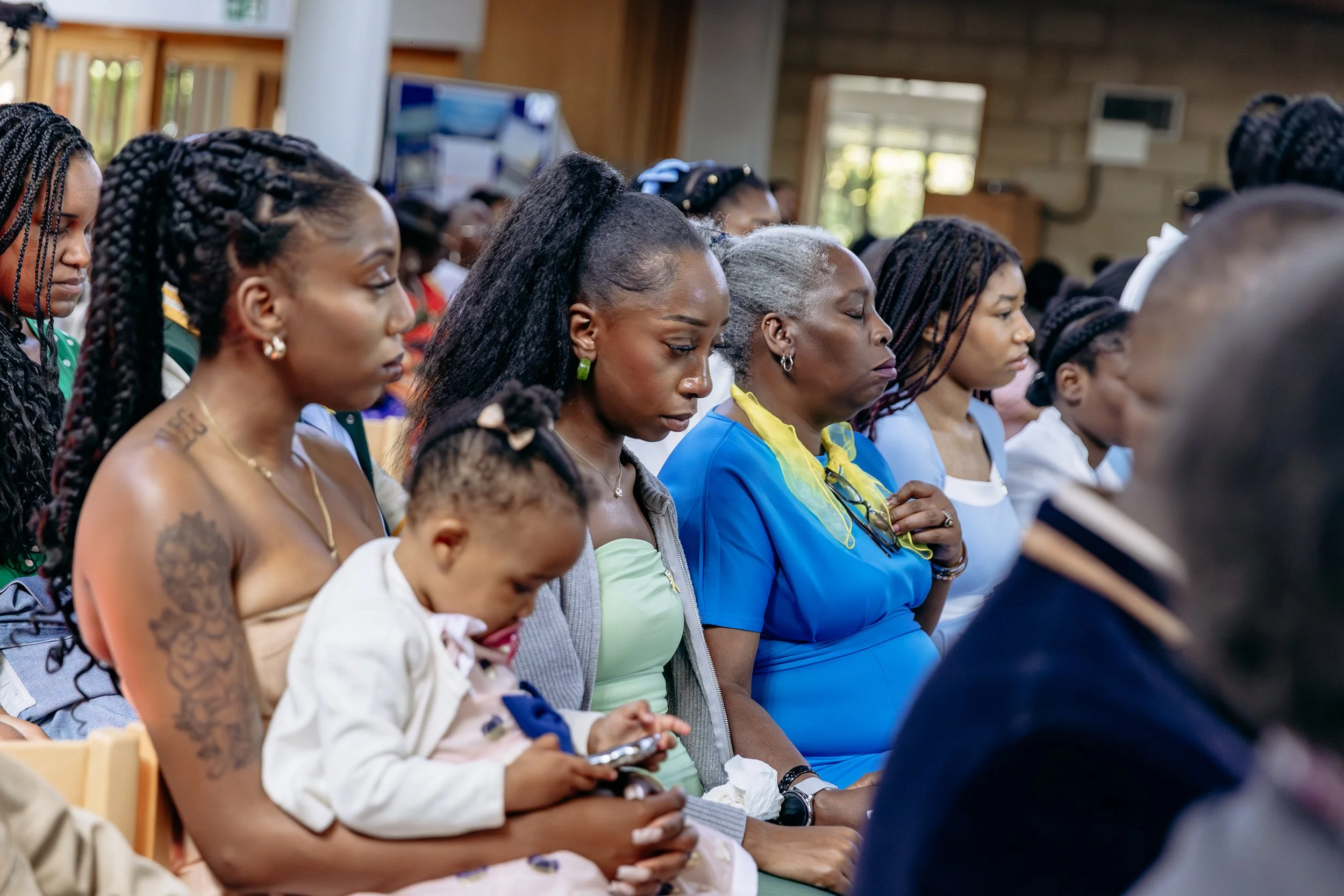 A group of women and a young girl sitting closely, attending a church or community gathering with their eyes closed in prayer.