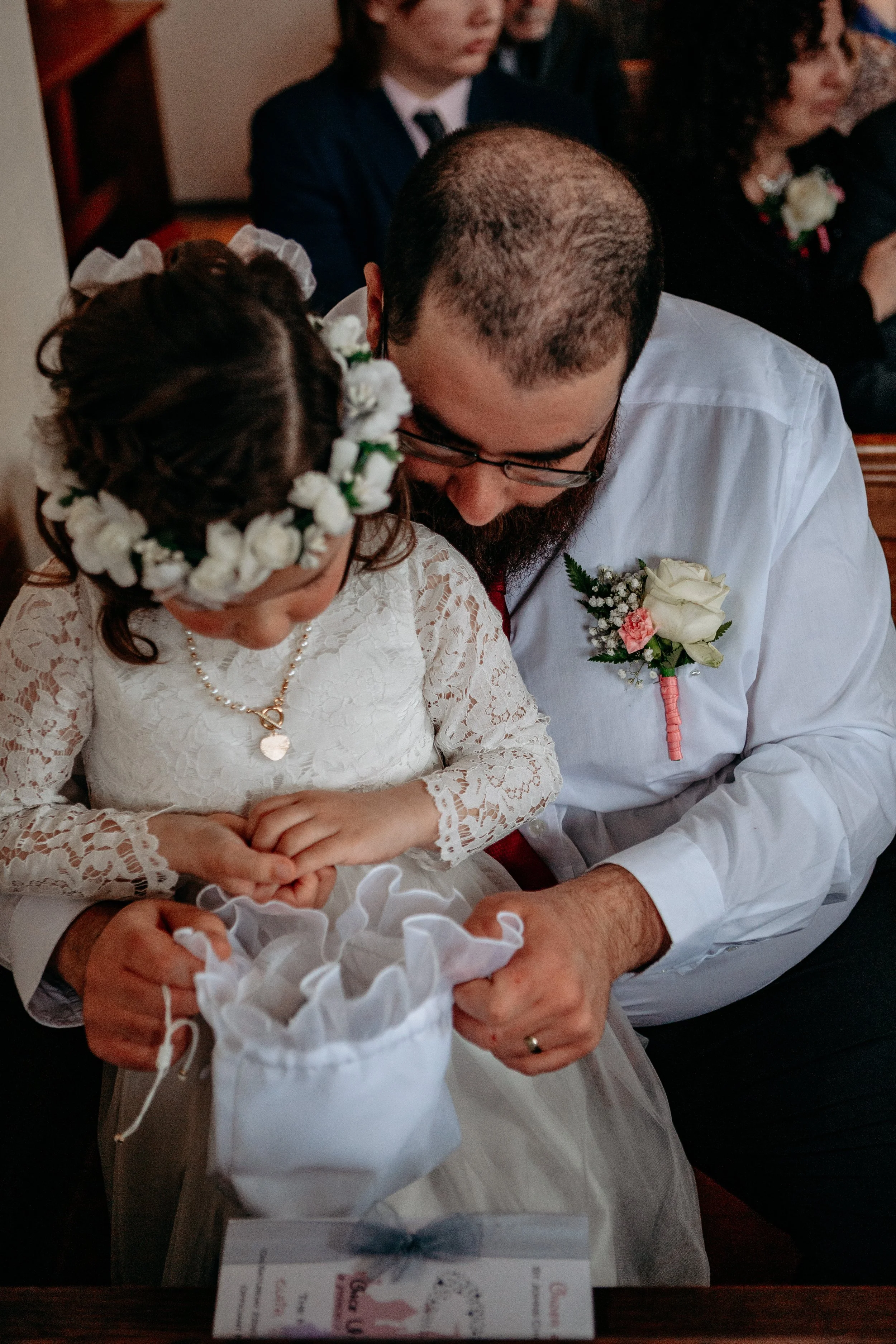 A young girl in a white dress with a floral crown looks into a small bag held by a man in a white shirt at a wedding ceremony.