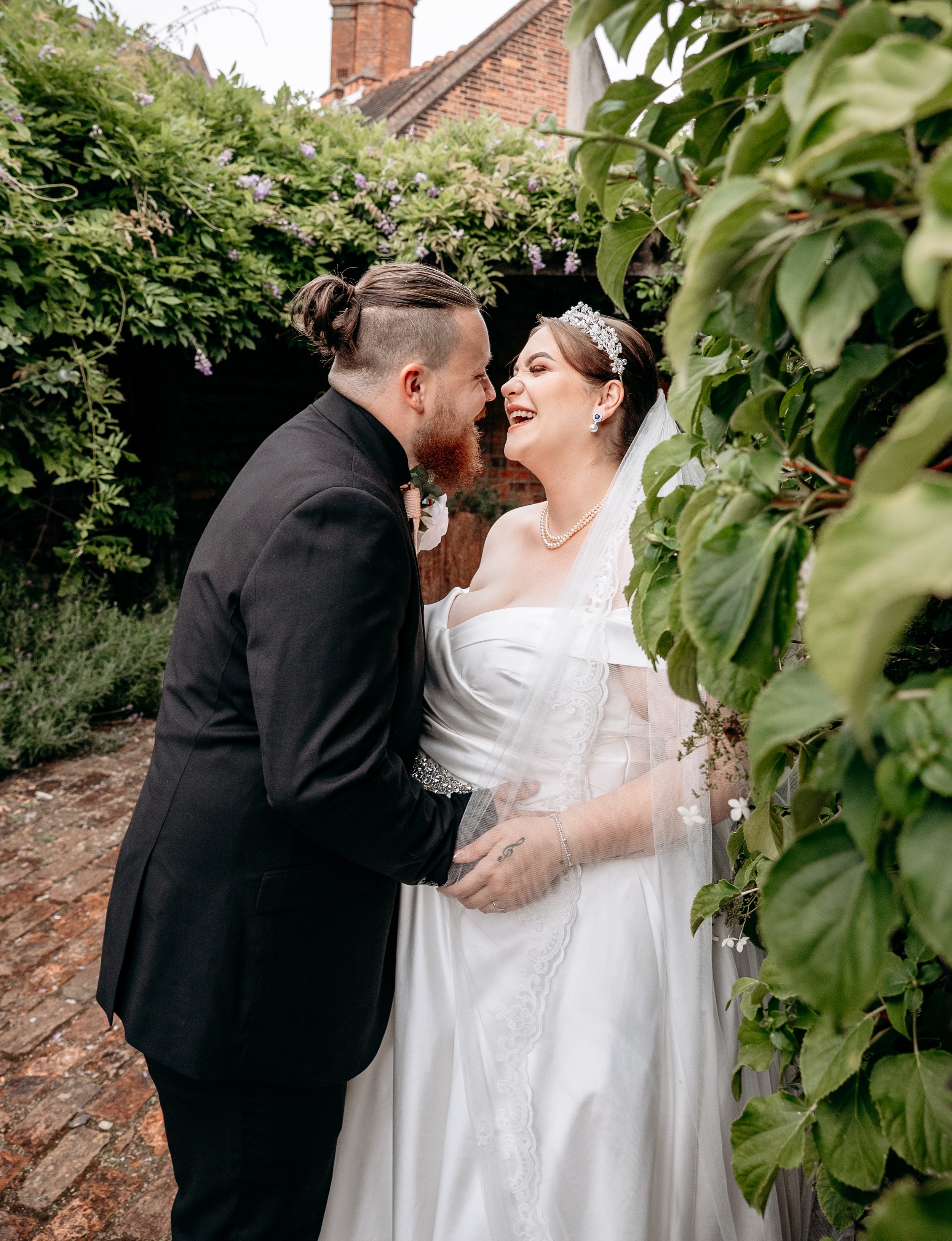 Bride and groom laughing closely together outdoors in a garden setting, with lush green foliage and brick pathways.