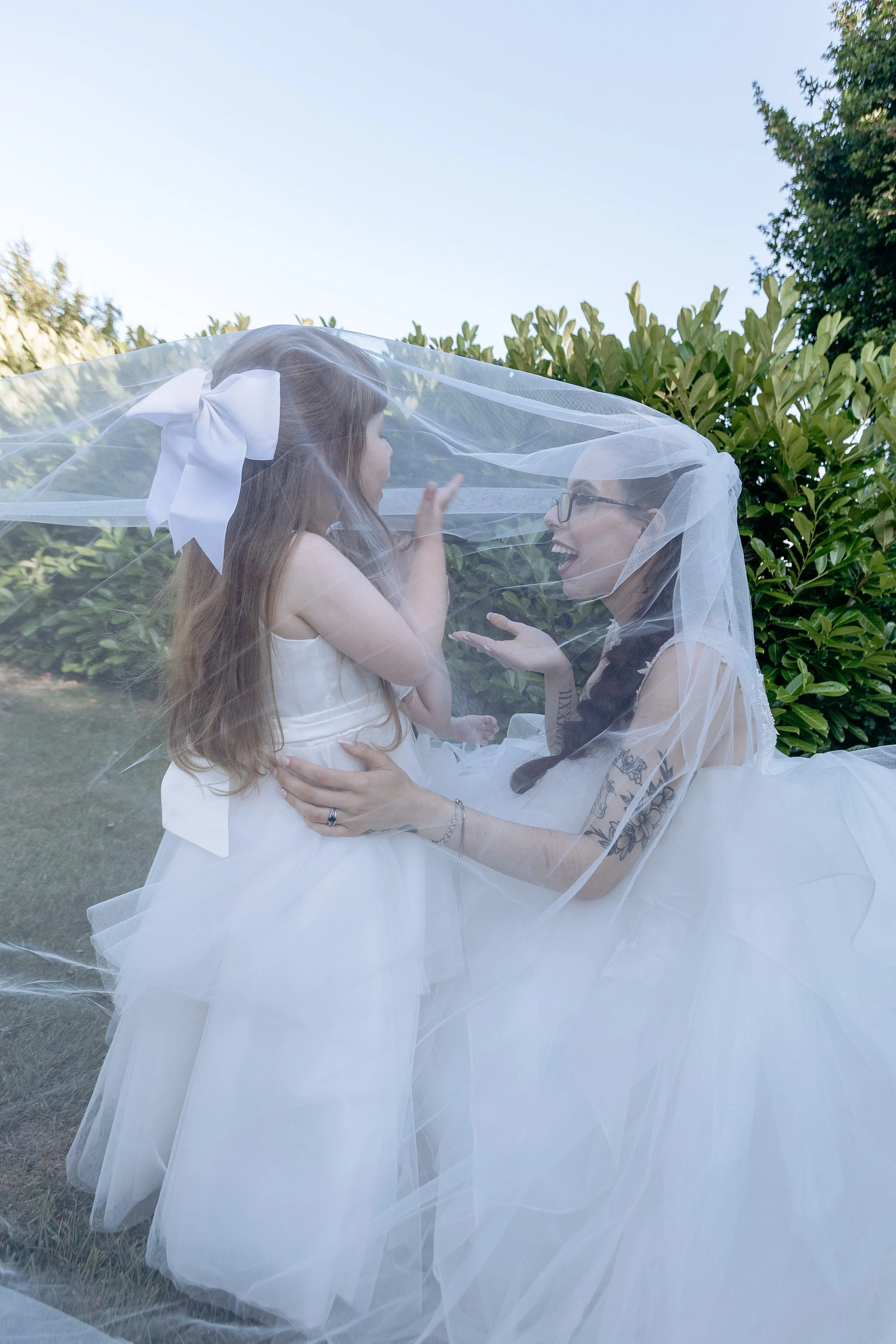 A woman and a girl dressed in white wedding gowns playing under a sheer veil outdoors, smiling and interacting with each other.