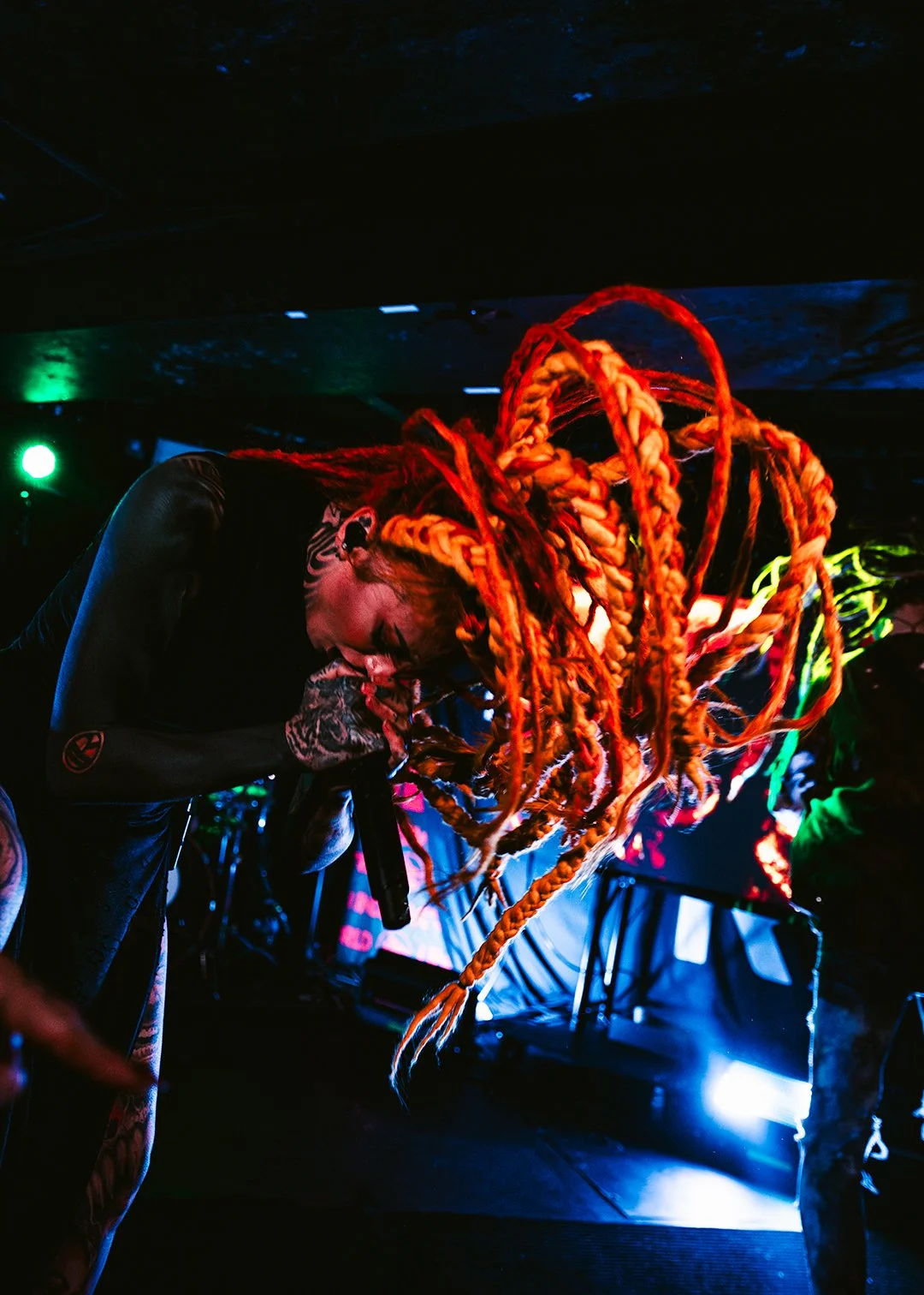 A performer with neon orange dreadlocks and tattoos, holding a microphone, on stage with colorful lighting.