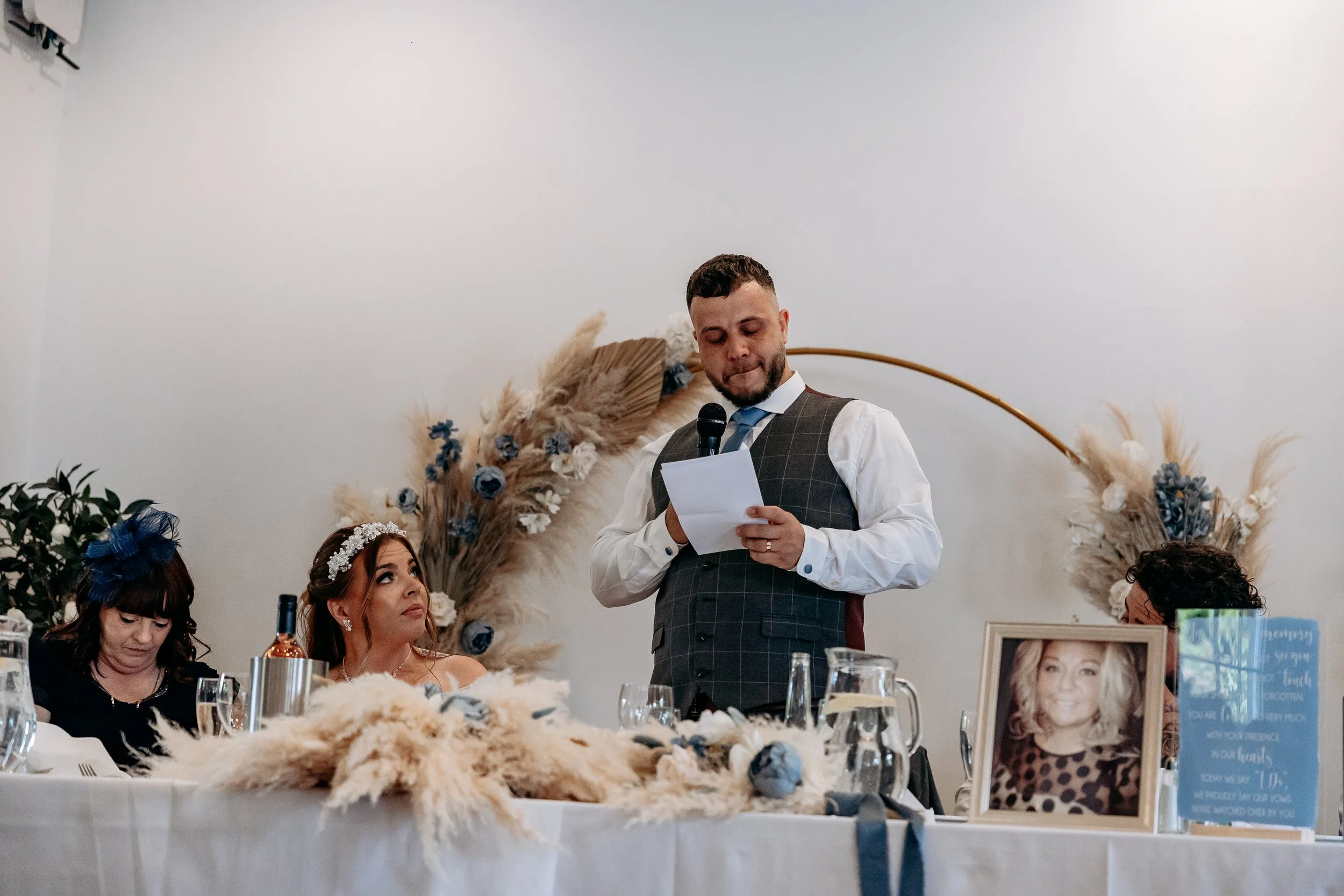 A man giving a speech at a wedding reception table, with a woman in a white dress and floral headband looking up at him, and two women sitting on either side of her.