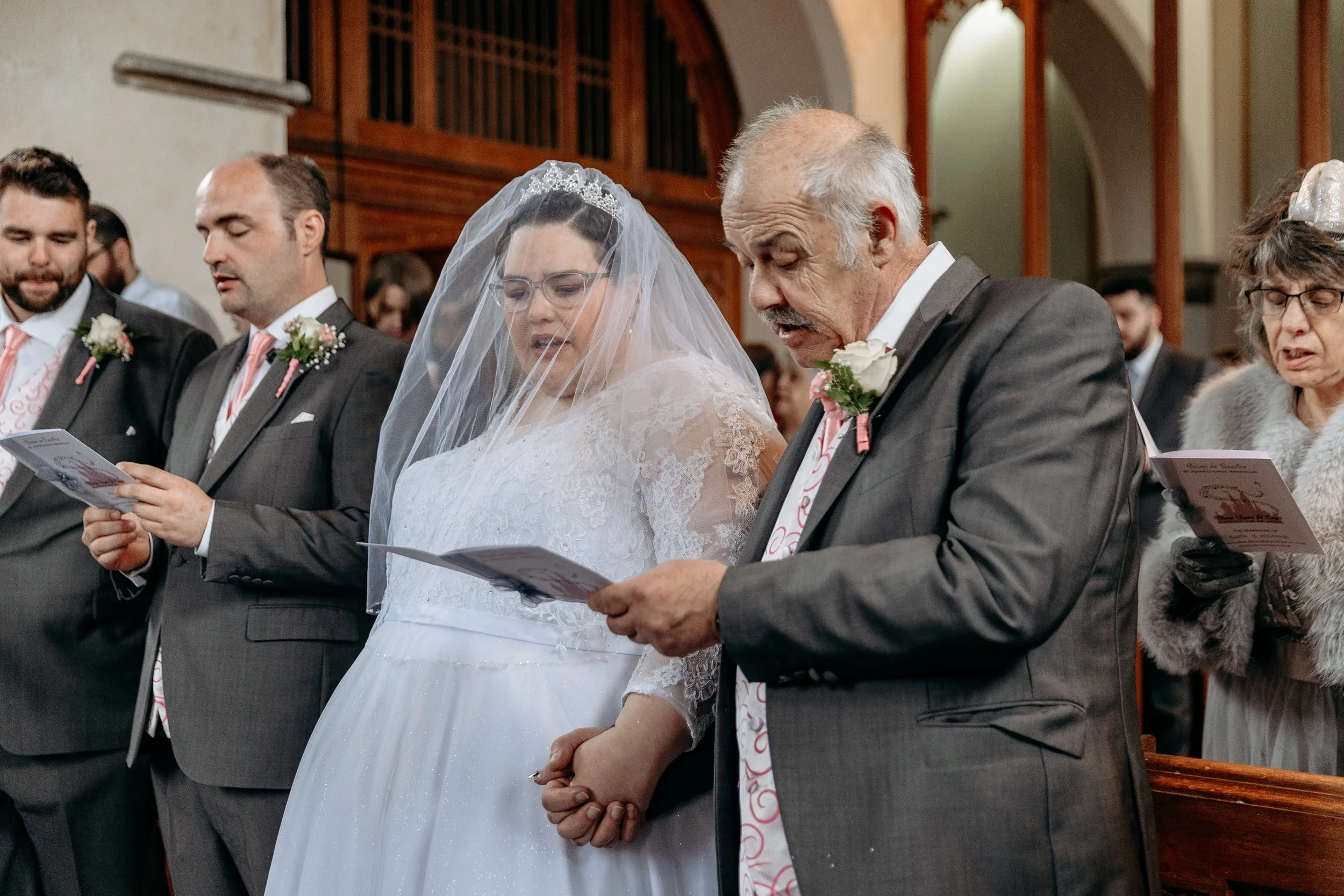 A bride and groom along with family members participating in a wedding ceremony inside a church, reading from booklets.
