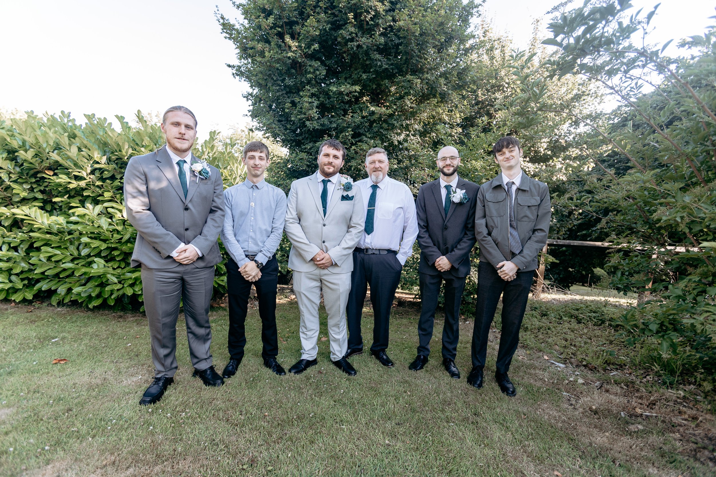 Group of six men in suits standing outdoors on grass with trees and bushes in the background, smiling at the camera.