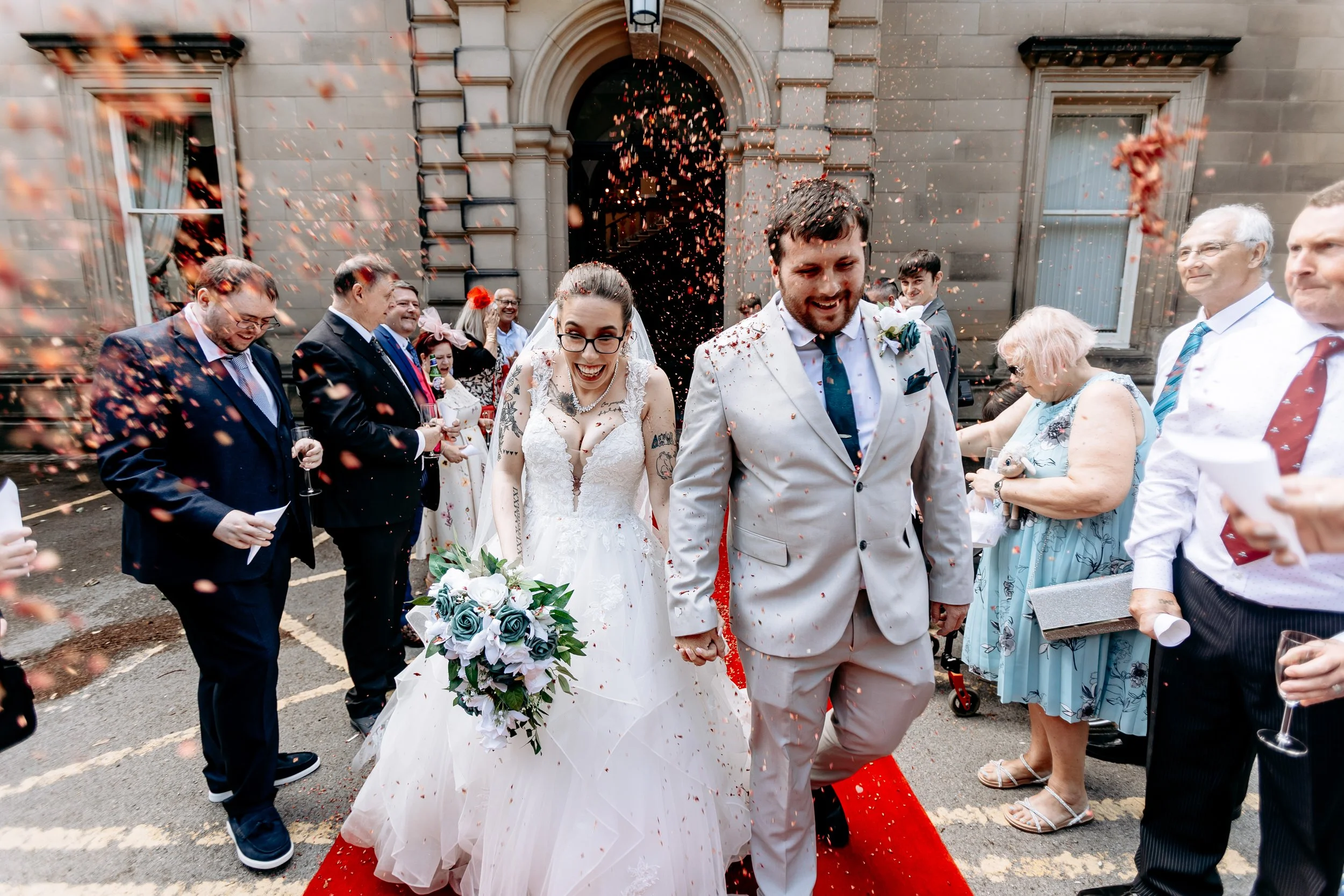 Bride and groom walking on a red carpet surrounded by guests throwing confetti during their wedding celebration outside a stone building.