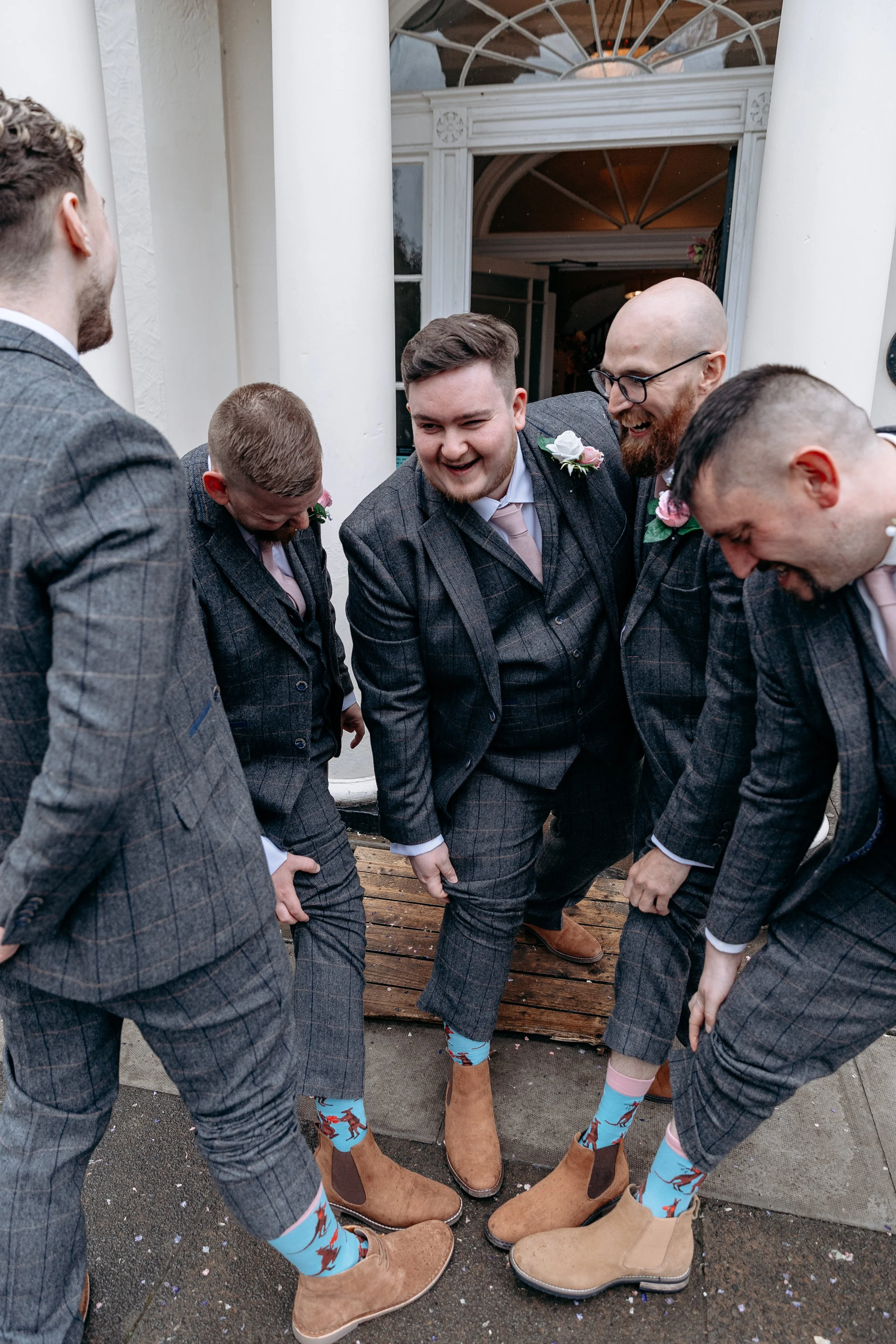 A group of men in checkered suits and colorful socks looking at their boots and smiling outside a building at a wedding.