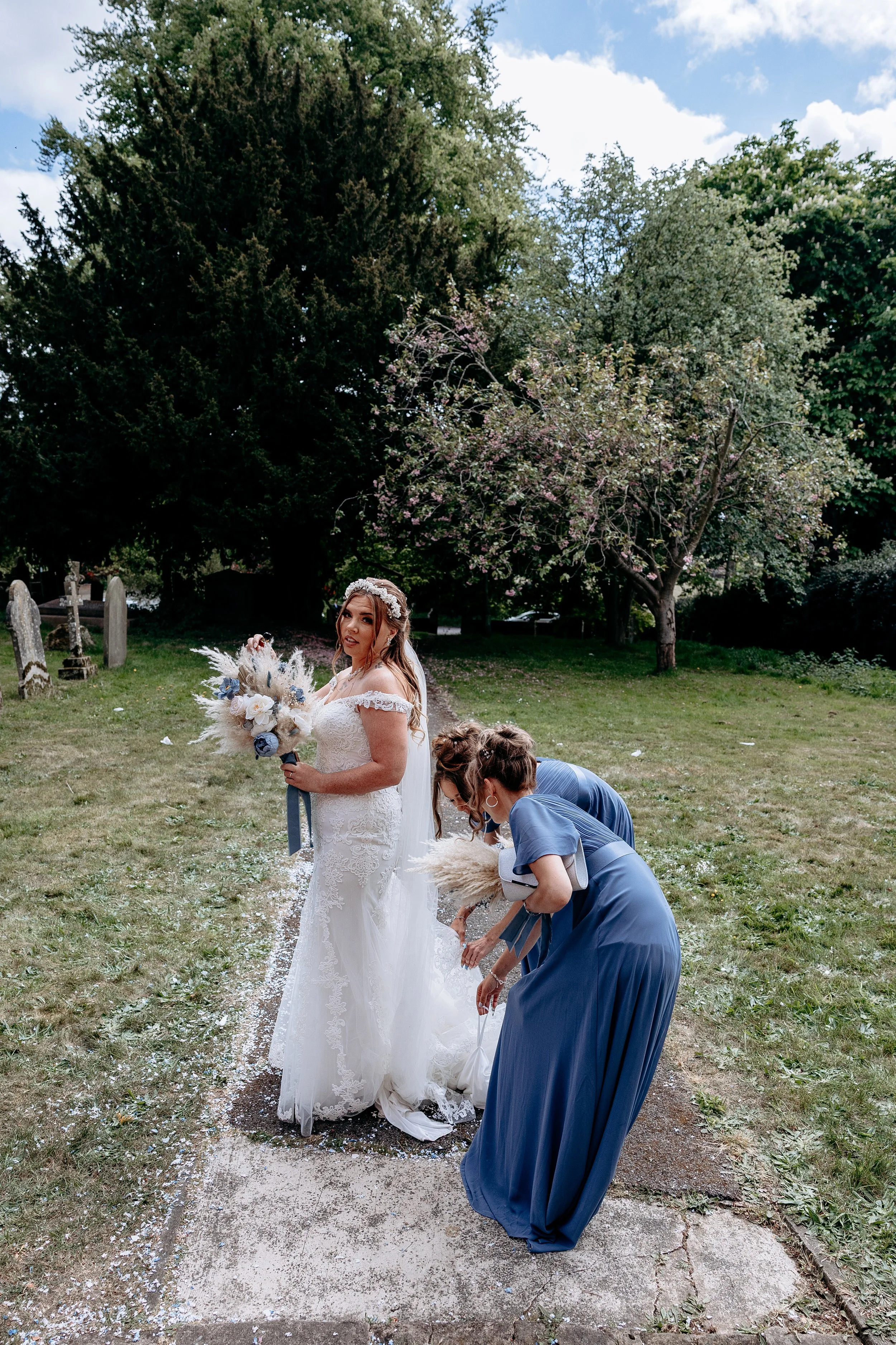 Bride in a white wedding dress holding a bouquet, with bridesmaids in blue dresses adjusting her train outdoors on a grassy area near trees.