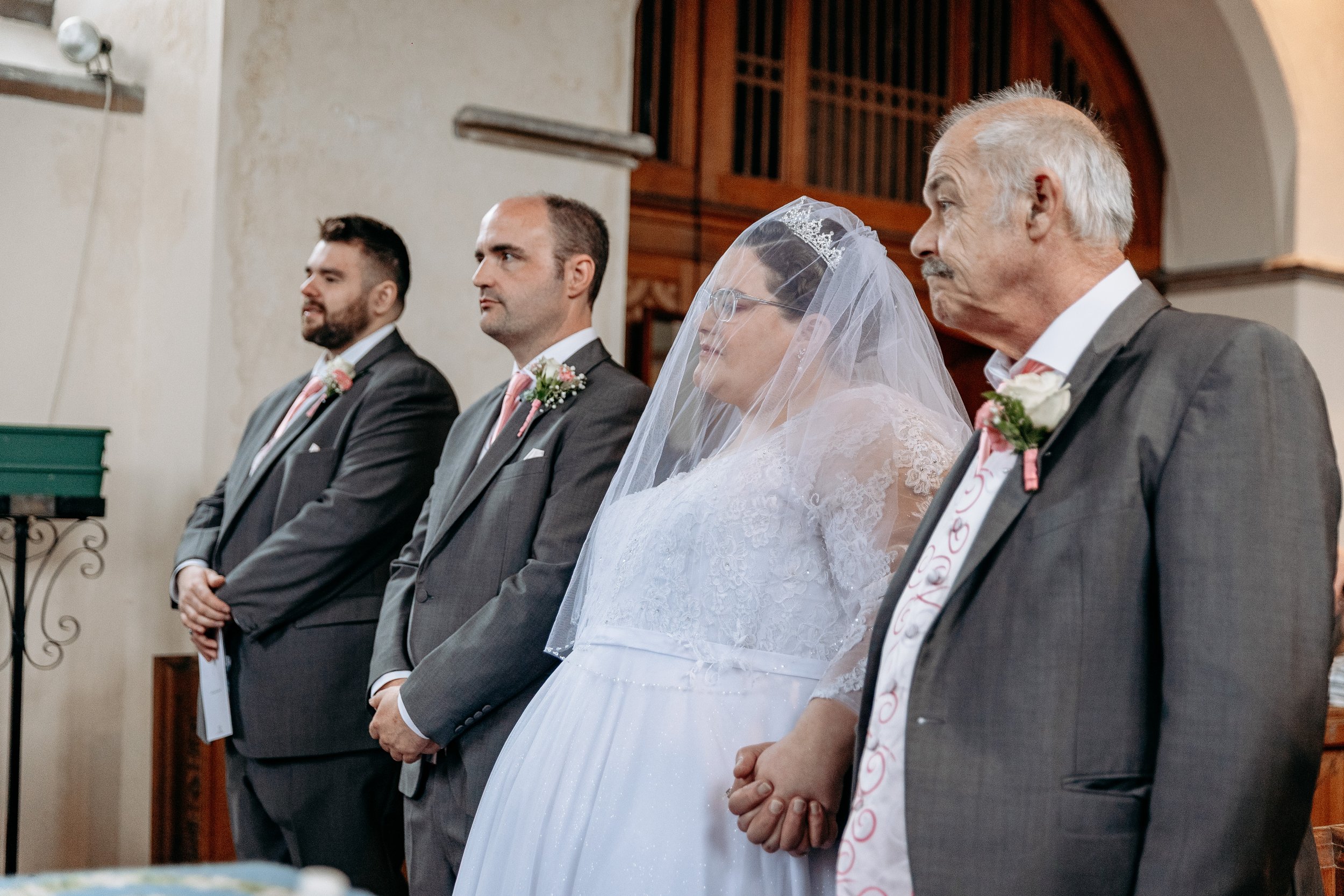 Bride in a white wedding dress and veil holding hands with an older man in a suit during a wedding ceremony, standing alongside three men in suits.