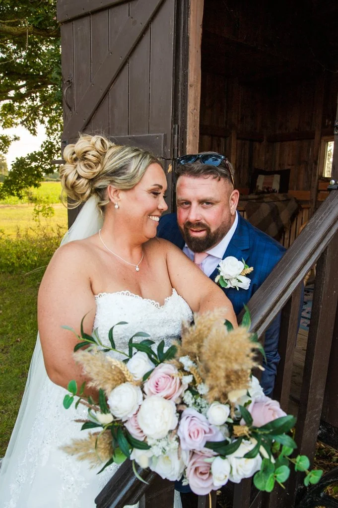Bride with a floral bouquet smiling with groom in a blue suit, inside a rustic wooden barn.