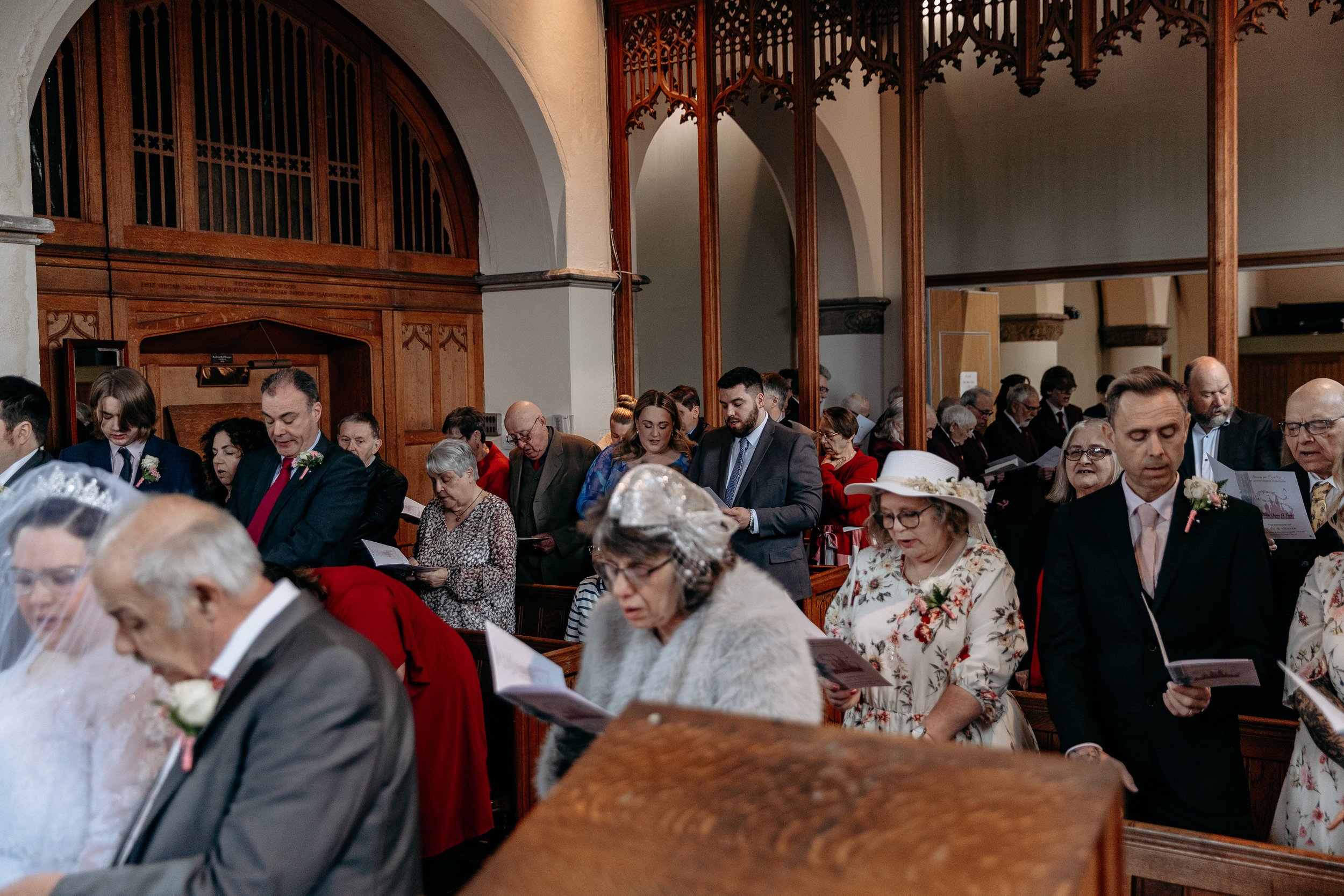 People attending a religious service or ceremony inside a church, standing and reading from booklets, with some dressed in formal attire and others in vintage or traditional clothing.