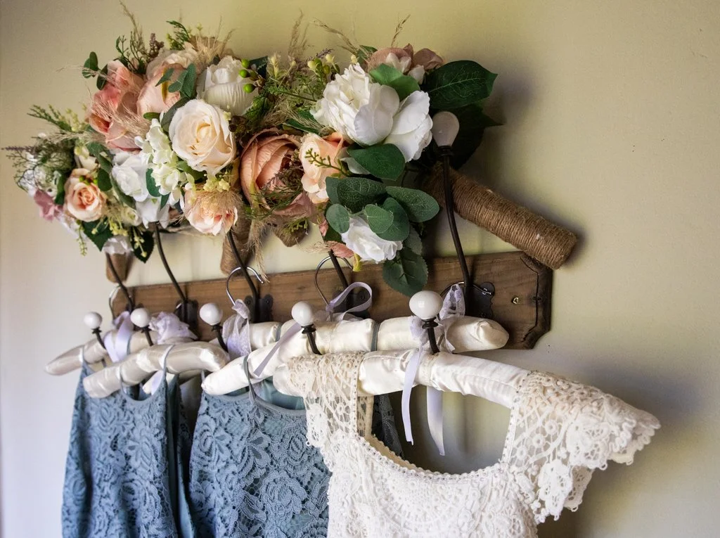 A wooden wall hook holding decorative hanging rods with lace dresses and a bouquet of pink, white, and cream flowers, accented with green leaves.