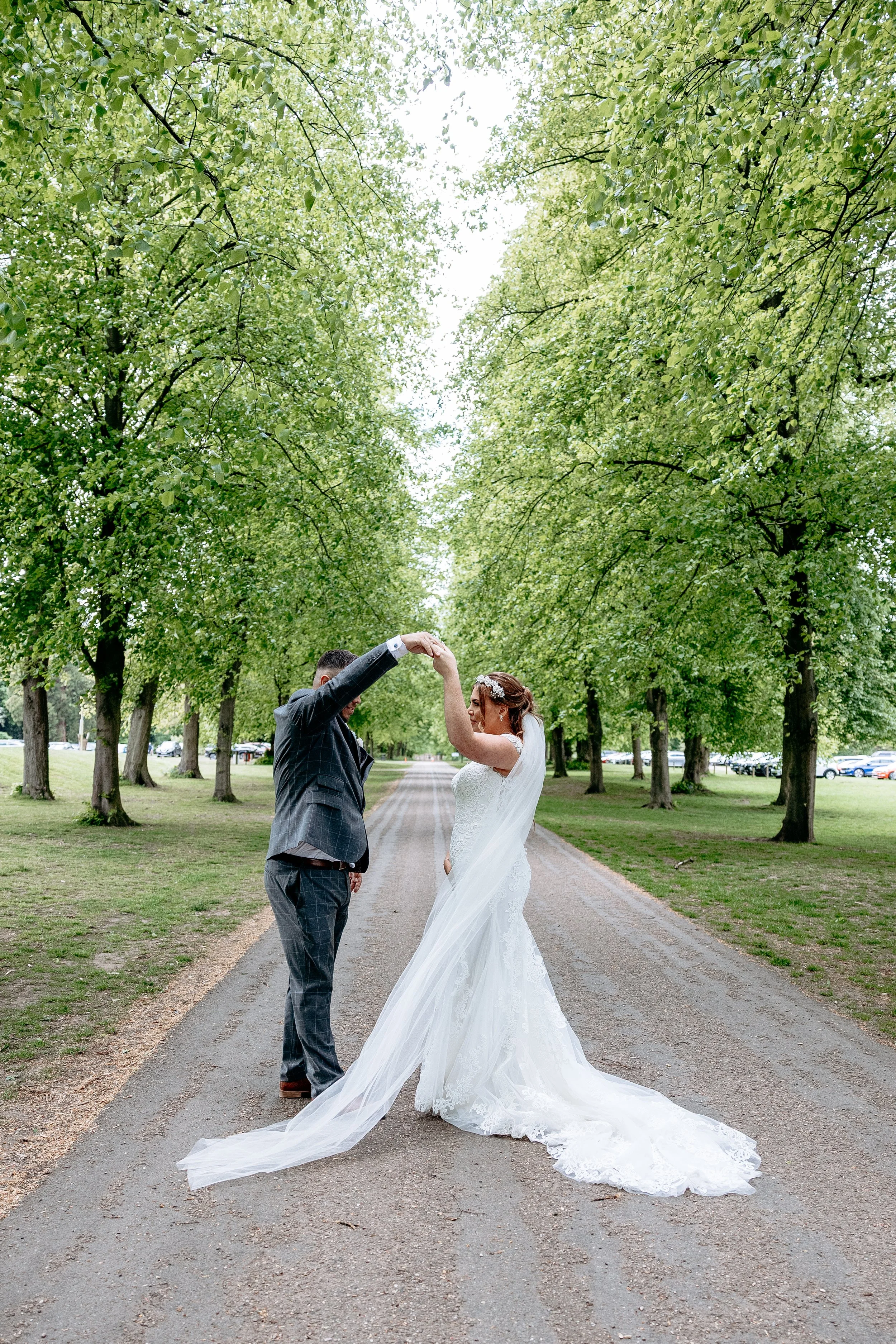 A bride and groom dancing on a tree-lined road during their wedding in the daytime.