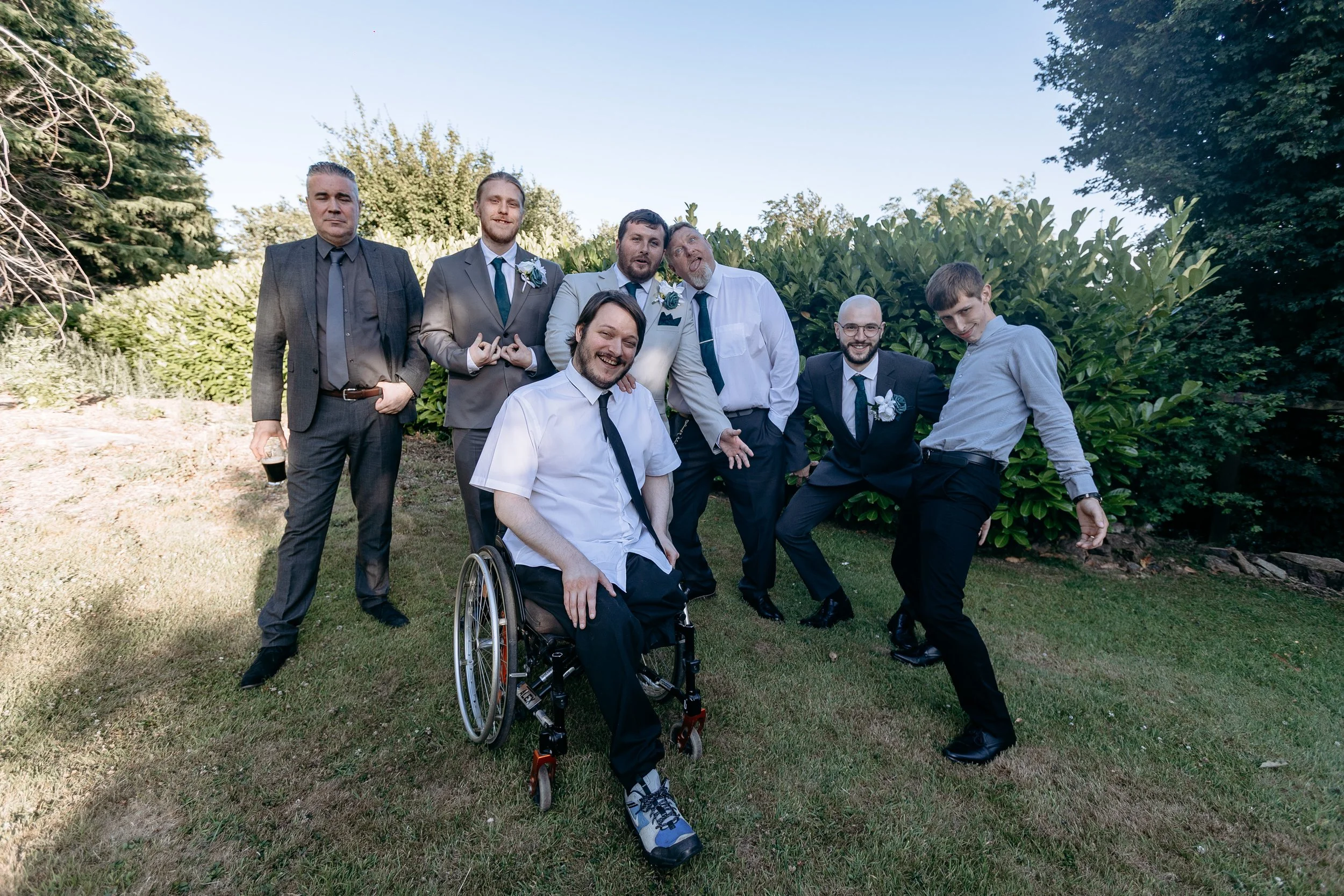A group of seven men, some in suits with boutonnieres and others in dress shirts, posing together outdoors on a grassy area with trees and bushes in the background. One man is in a wheelchair, and all are smiling or making playful gestures.