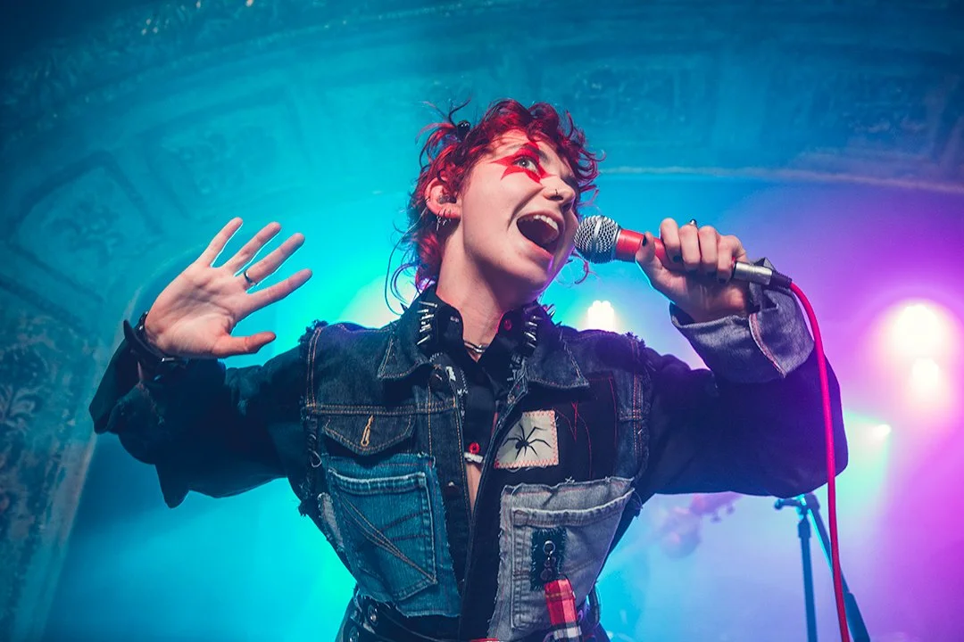 A woman with red hair, star face paint, and a denim jacket singing into a microphone on stage with colorful lights.