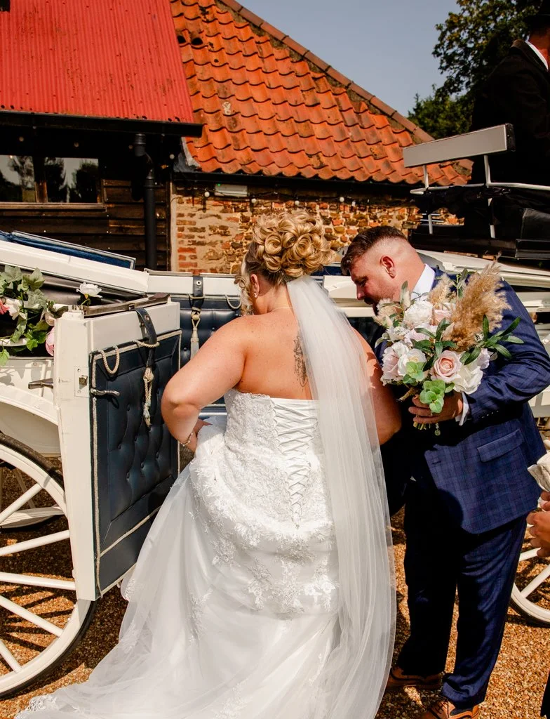 Bride in a white wedding dress and veil standing next to a horse-drawn carriage, with groom in a navy suit holding a bouquet of flowers, during a wedding ceremony outside near a rustic brick and wood building.