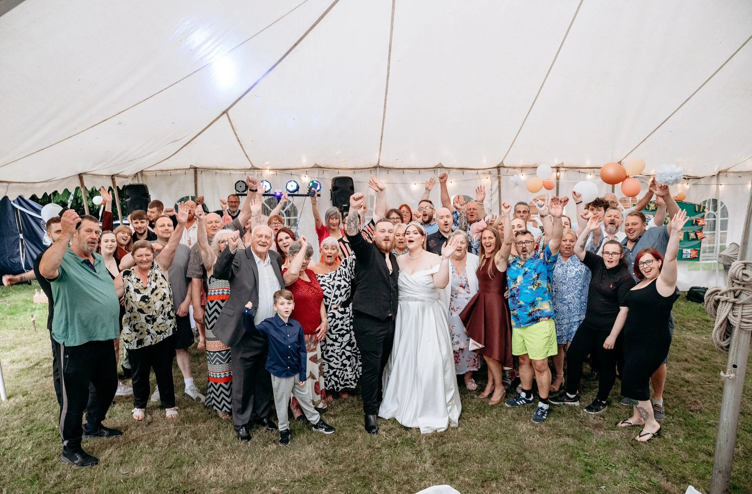 Large group of people celebrating inside a decorated tent, with many raising their hands and smiling, including a bride and groom in the center.