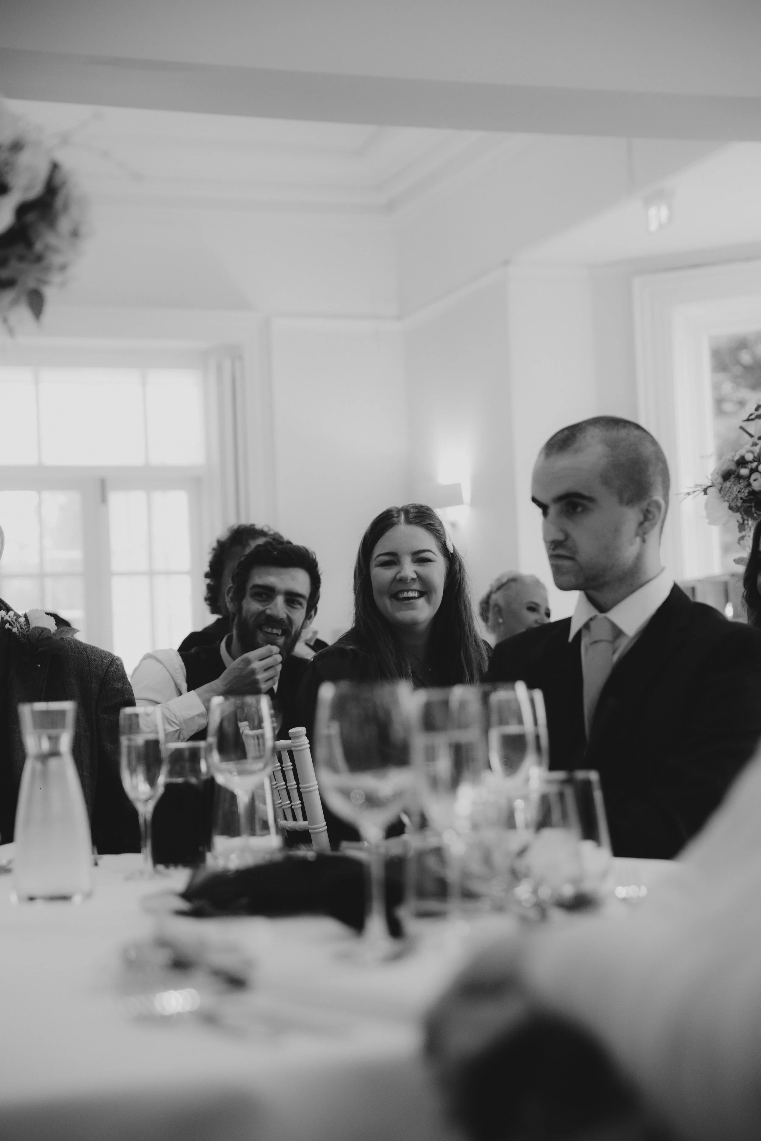 A group of people sitting at a table during a celebration, smiling and talking, in a well-lit room with large windows.