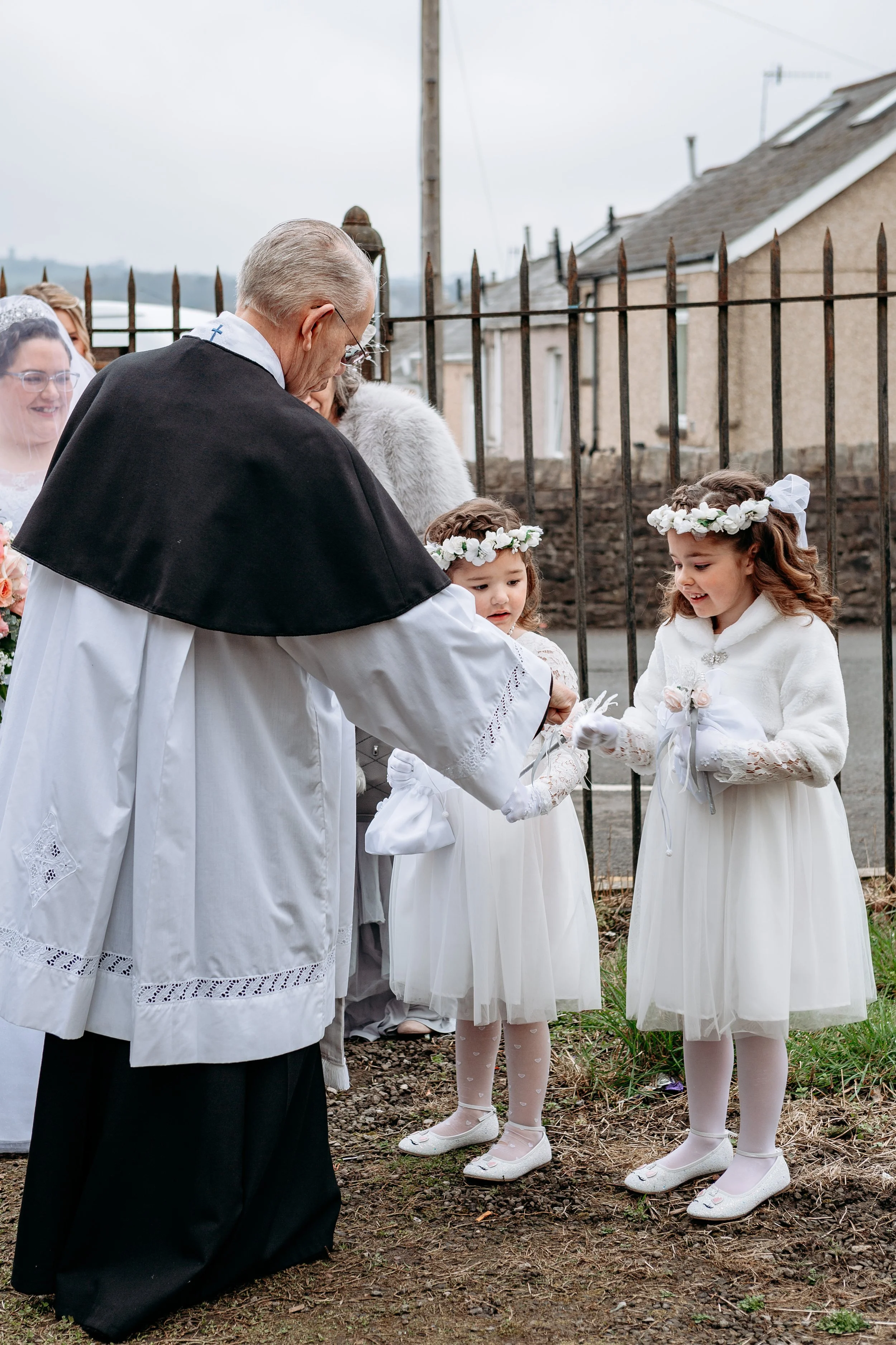 A priest conducting a First Communion ceremony outdoors, with two young girls dressed in white, wearing flower crowns, receiving a blessing from the priest.